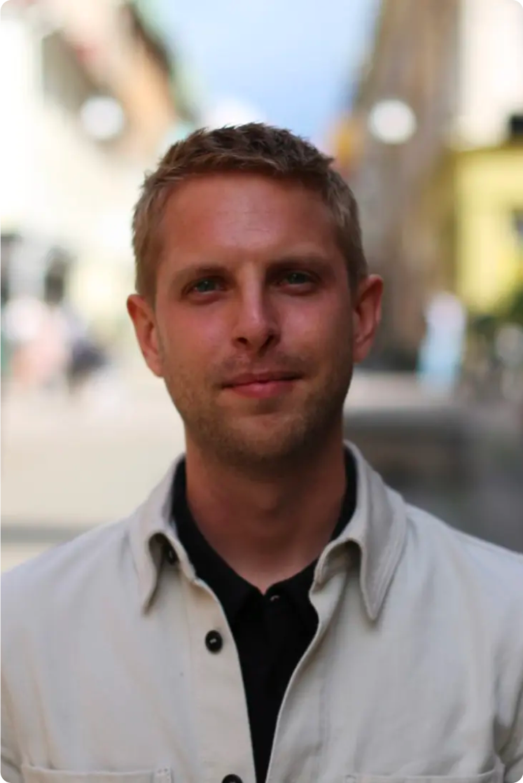 A young man with light brown hair and blue eyes standing outdoors with blurred buildings and trees in the background.