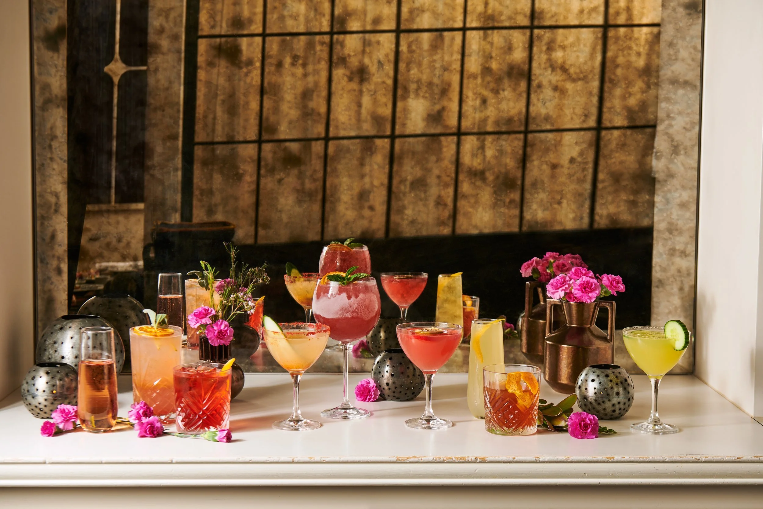 An assortment of colorful cocktails and drinks displayed on a white surface with pink flowers, black decorative objects, and a mirror background.
