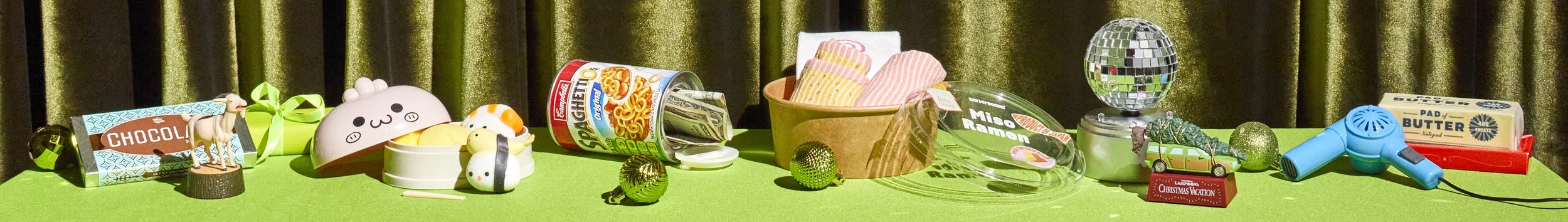 A collection of assorted household and decorative items on a bright green surface with a background of brown curtains, including toy figures, a can of instant noodles, a bowl of socks, a small toy car, a disco ball, a hairdryer, and boxes of butter.