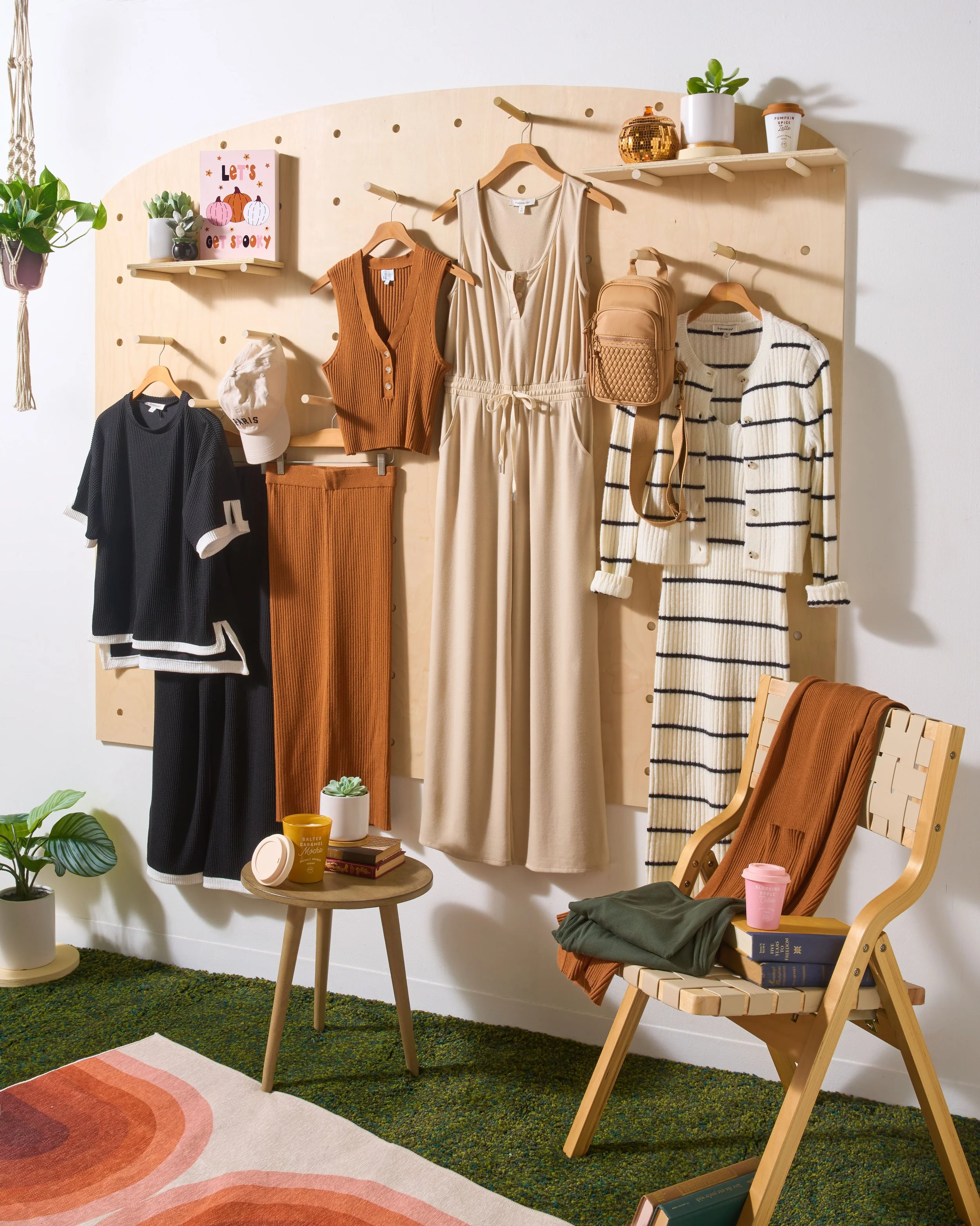 A wall display of hanging clothing and accessories in a cozy room. The display includes a beige jumpsuit, a striped cardigan, a black and white shirt, a brown vest, and a pair of brown pants. There are also hats, a backpack, and potted plants. A smal