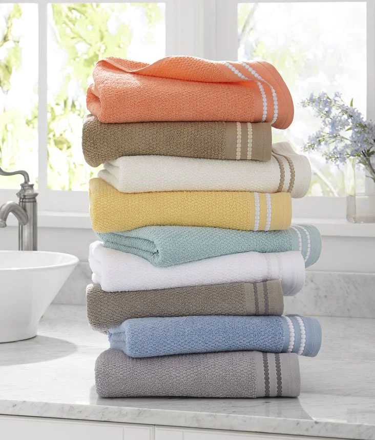 Stack of folded towels in various colors on a white marble bathroom counter with a window and a flower vase in the background.