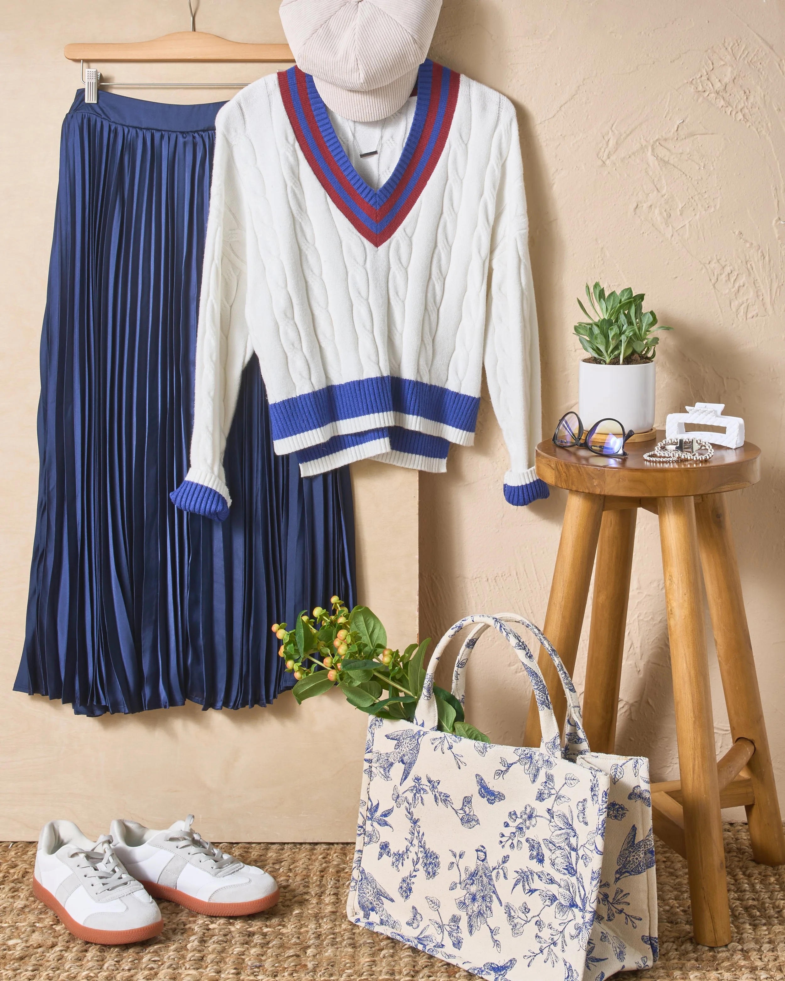A pair of white sneakers with a gum sole, a blue pleated skirt hanging on a hanger, a white cable knit sweater with blue and red accents, a beige hooded top, a potted plant, sunglasses, jewelry, a small table, a tote bag with blue floral pattern, and