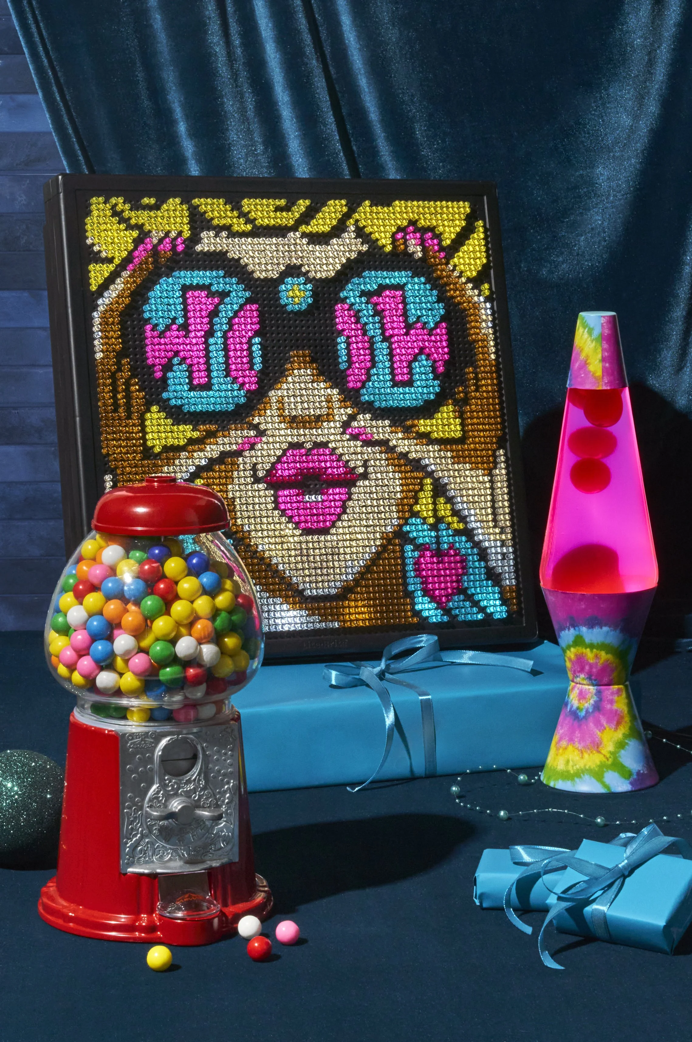 Decorative display with a colorful piece of artwork, a gumball machine filled with multicolored gumballs, a pink tie-dye lava lamp, and wrapped presents on a dark table with a blue curtain background.