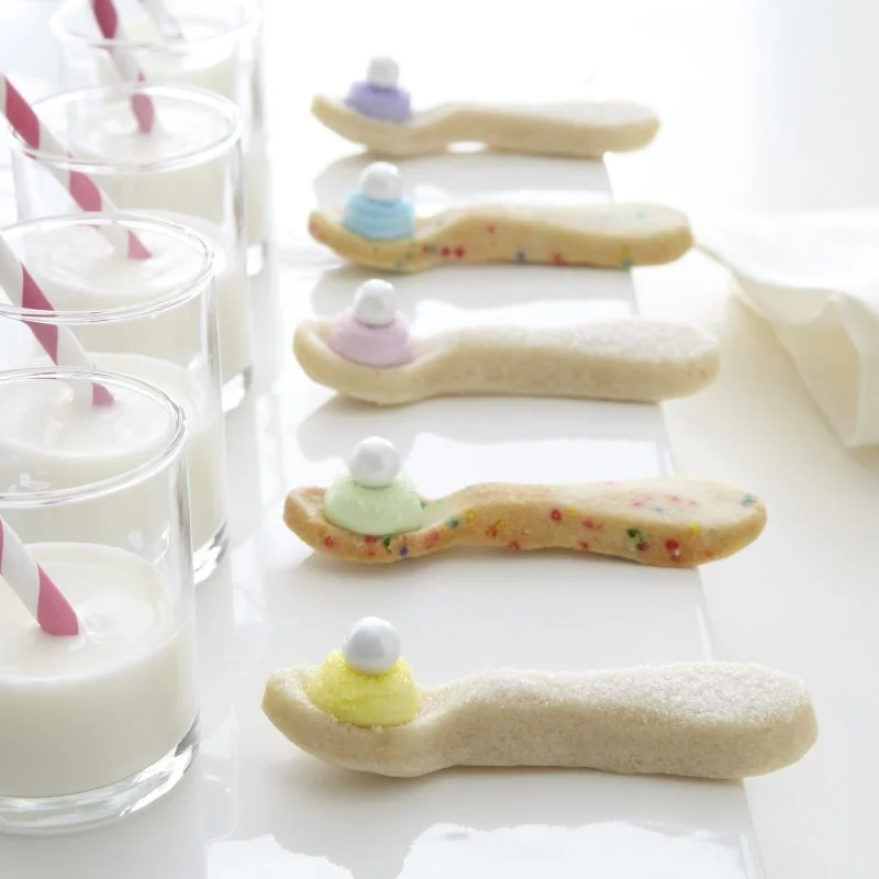 Five ice cream spoon cookies decorated with pastel-colored frosting and sprinkles, lined up next to glasses of white milk with pink and white striped straws.