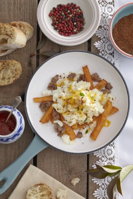 A breakfast plate with mashed potatoes, meat, and fried sweet potato fries, along with slices of bread, a bowl of mixed berries, and a cup of coffee on a wooden table with a lace doily.