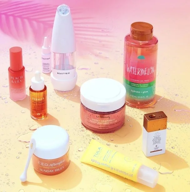 Collection of skincare and beauty products, including serums, creams, gel, and a facial device, arranged on a pink and yellow surface with water droplets.
