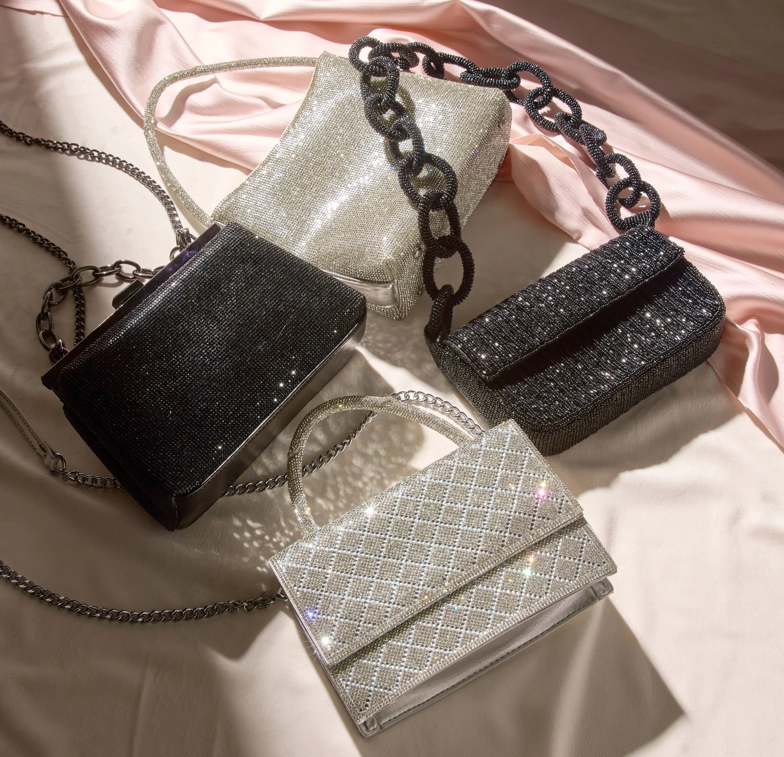 Five shiny handbags with chain straps arranged on a bed with beige and pink satin fabric.