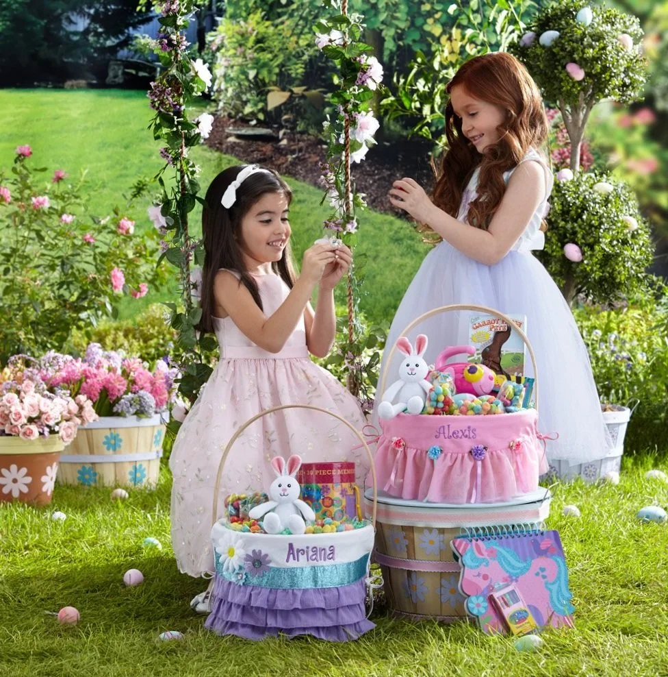 Two young girls in dresses at an outdoor Easter egg hunt, surrounded by baskets of filled eggs and decorated with flowers and greenery.
