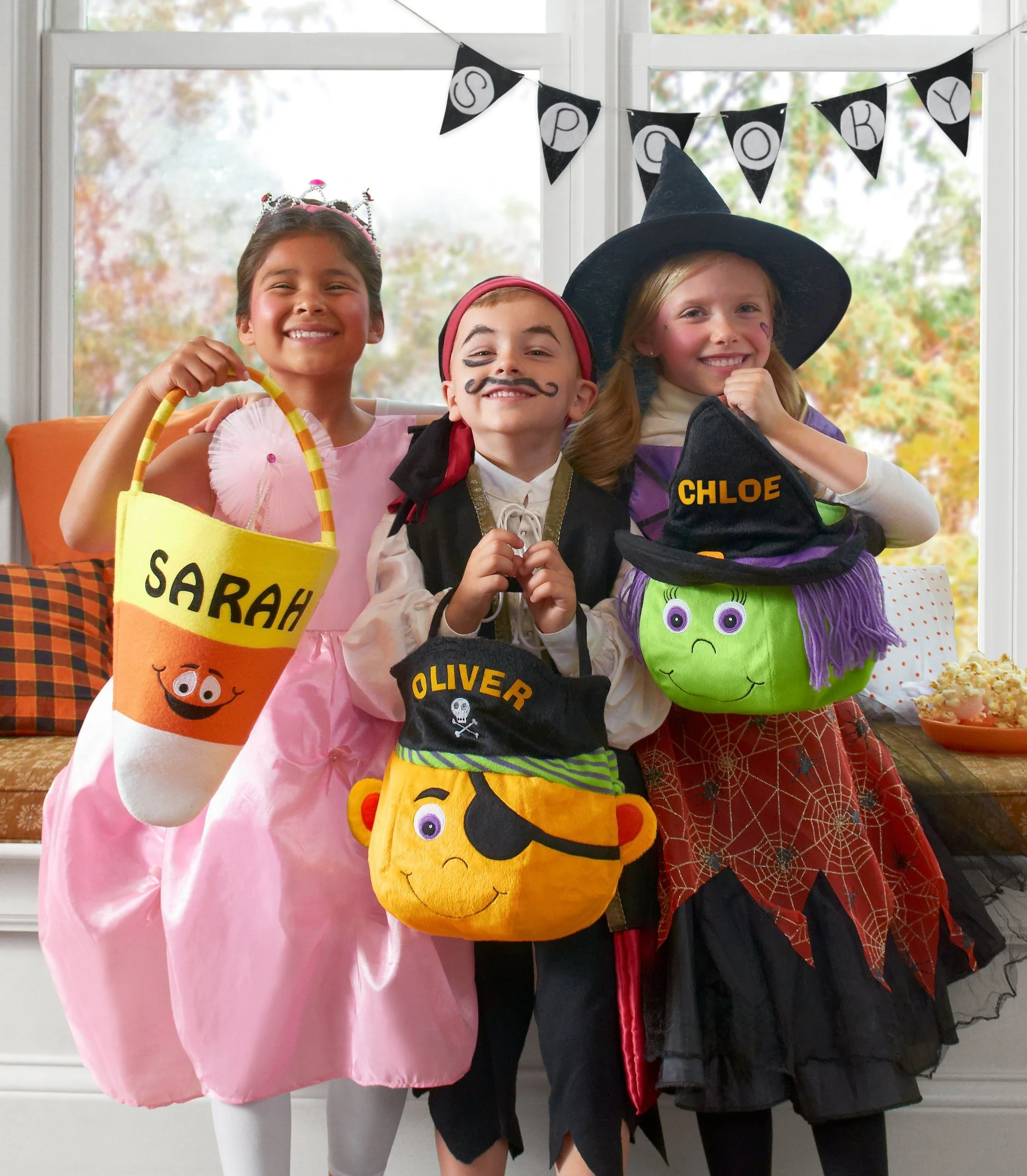 Three children dressed in Halloween costumes smiling and holding pumpkin-shaped mini backpacks, standing in a decorated room with a Halloween banner and fall foliage outside the window.