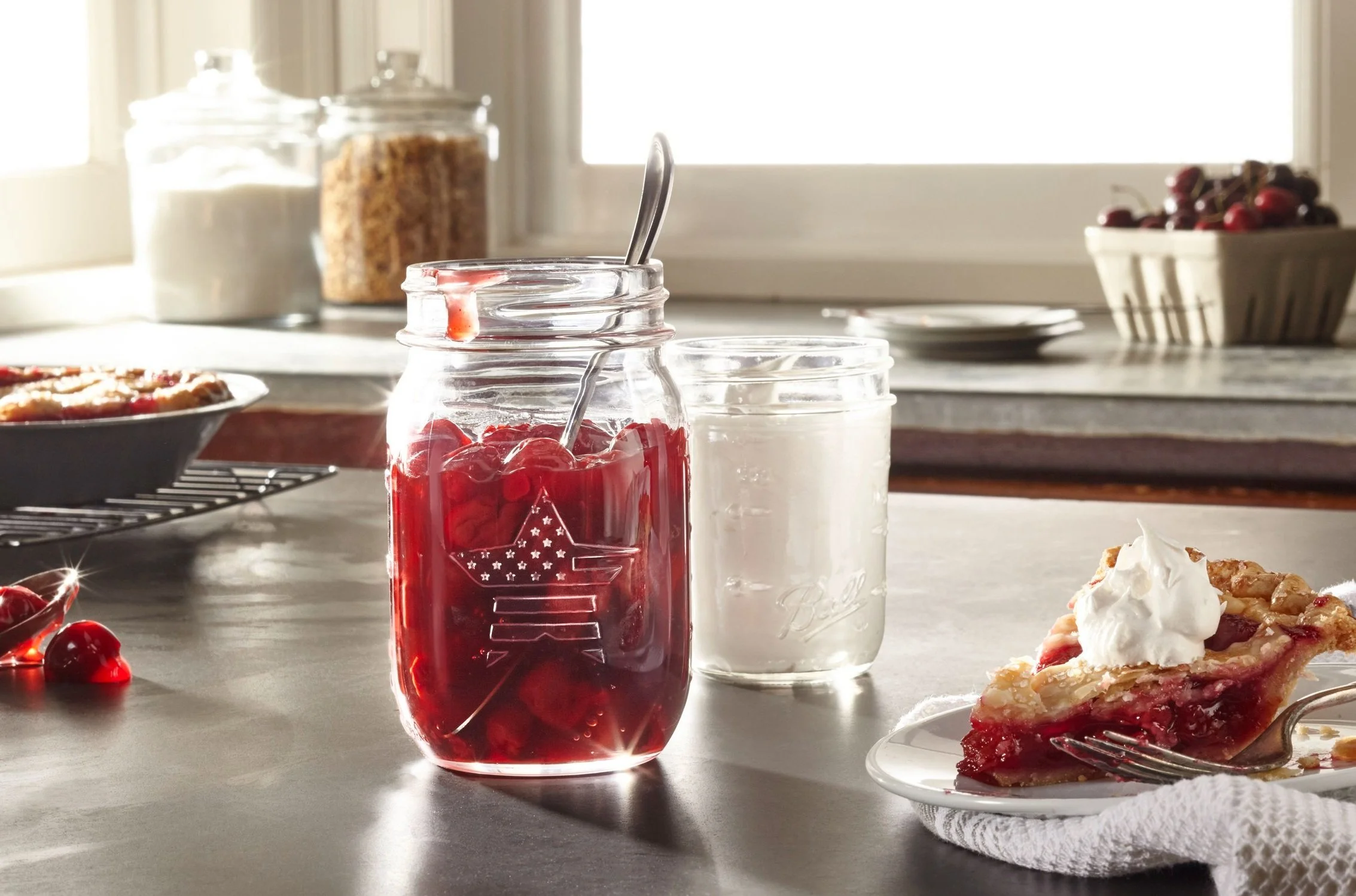 A slice of cherry pie with whipped cream on a white plate, a jar of cherry filling, and a glass of milk on a kitchen countertop, with a window in the background.