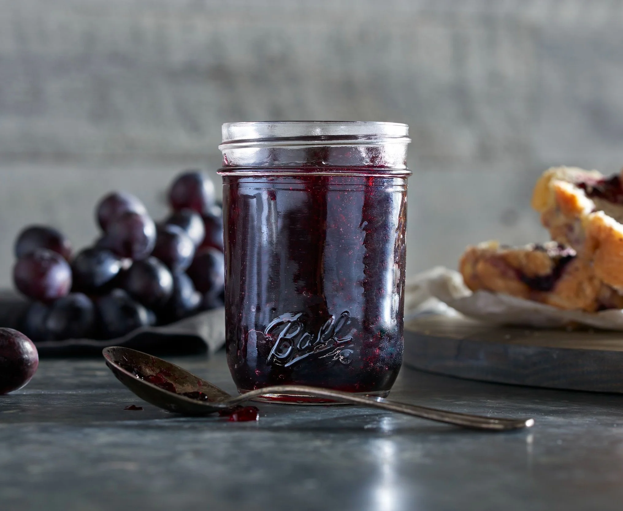 A jar of dark purple grape jam on a wooden surface with fresh grapes in the background and a partially visible plate of baked good to the right.