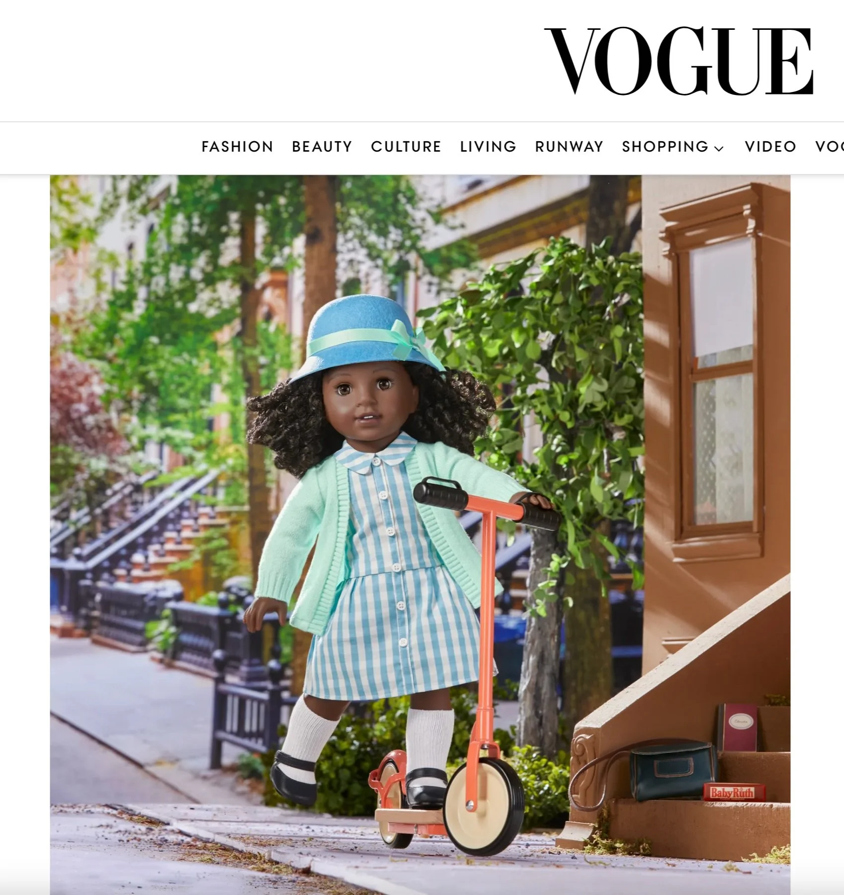 A doll with dark skin, curly black hair, wearing a blue hat with a ribbon, a light green sweater, a blue and white striped dress, white knee-high socks, and black shoes, riding a riding toy scooter on a sidewalk in an outdoor urban setting with green