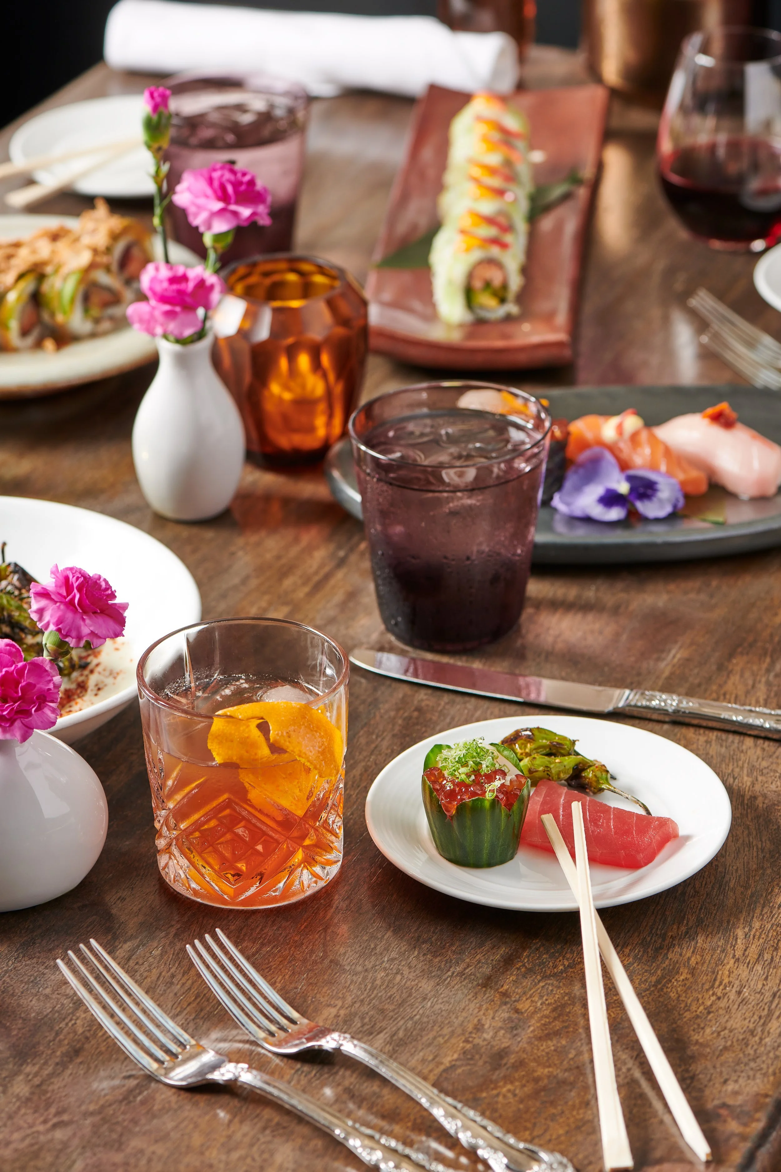 A dinner table set with various dishes, drinks, and flowers. The table features sushi rolls on a rectangular plate, sashimi with edible flowers on a black plate, a cocktail with orange peel in a patterned glass, a glass of water with ice, and a small