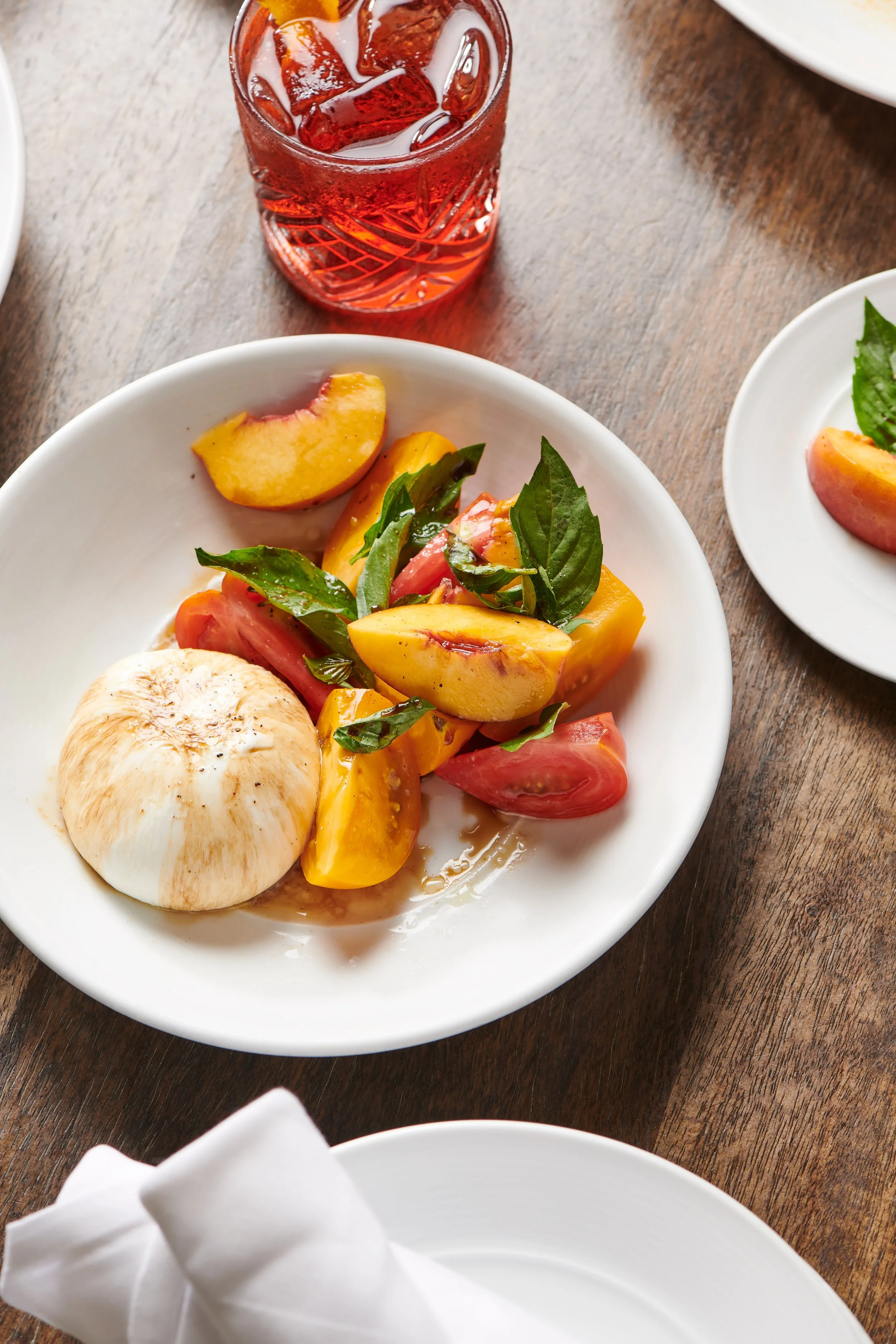 Caprese salad with fresh basil, heirloom tomatoes, and burrata cheese on a white plate, accompanied by a glass of red-colored drink with ice on a wooden table.