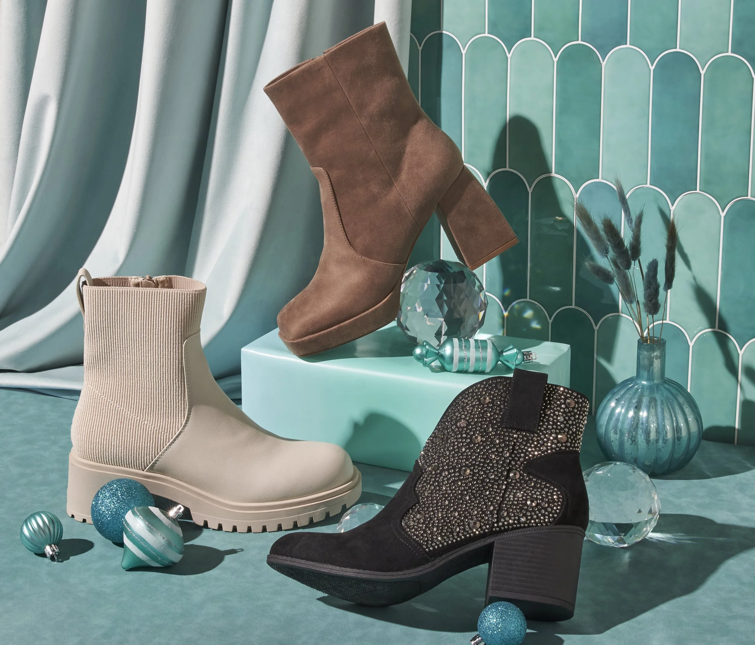 Display of three pairs of ankle boots with decorative ornaments and a vase with dried grasses, set against teal and blue patterned background with teal drapes.