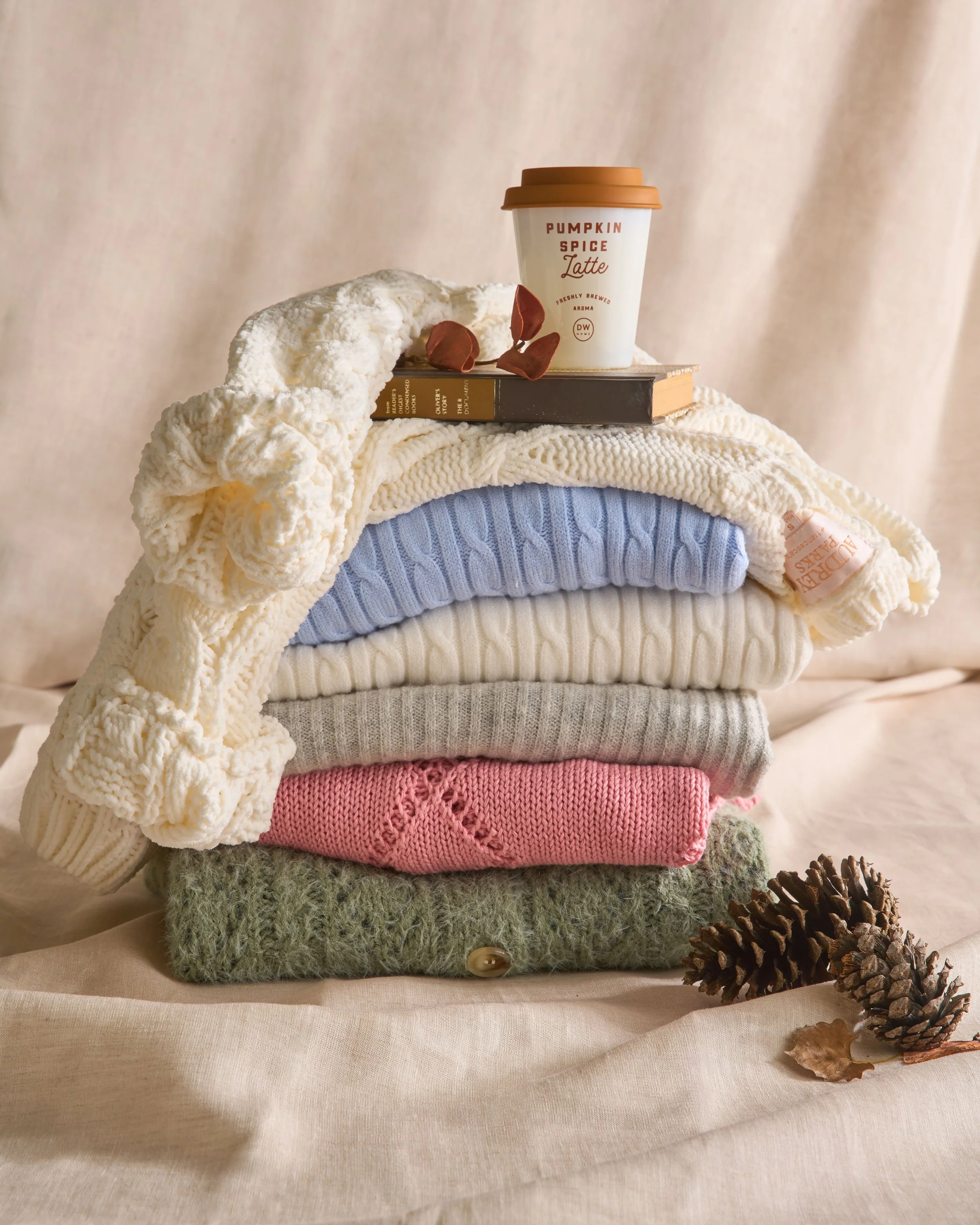 A stack of folded sweaters in different colors and textures, topped with a book and a cup of pumpkin spice latte, with pinecones and a dried leaf on a soft beige surface in front of a neutral background.