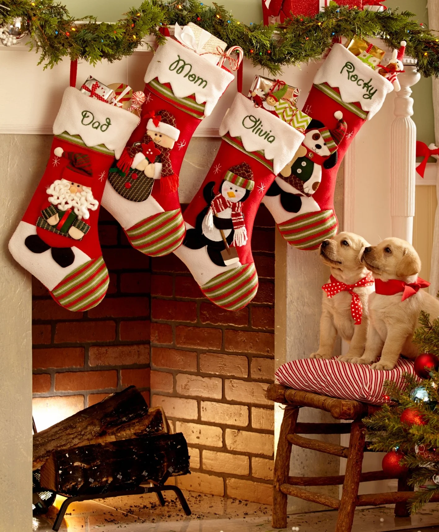 Festive Christmas scene with four red stockings hanging from a green garland over a brick fireplace, each labeled with names: Dad, Mom, Olivia, Rocky. The stockings are decorated with snowmen, Santa, and penguin figures. Two puppies with red bandanas