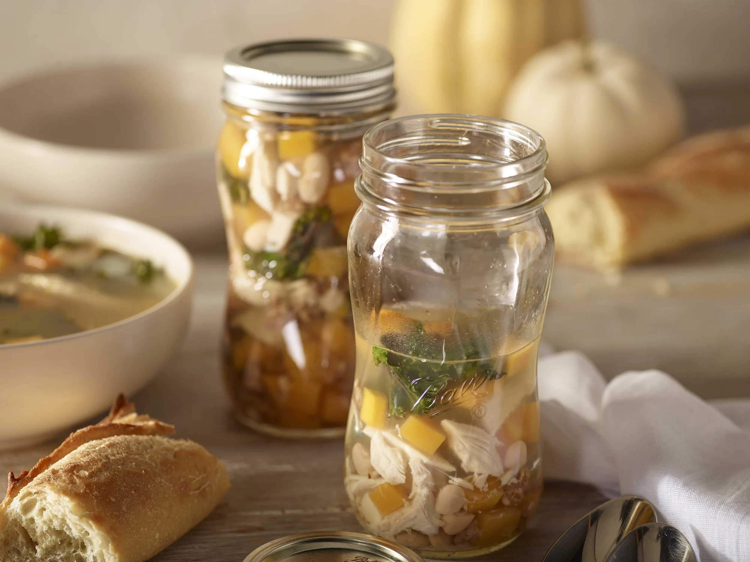 Two glass jars filled with a layered salad containing chicken, mango, spinach, and beans on a wooden table, with bread, a bowl of soup, and a pumpkin in the background.