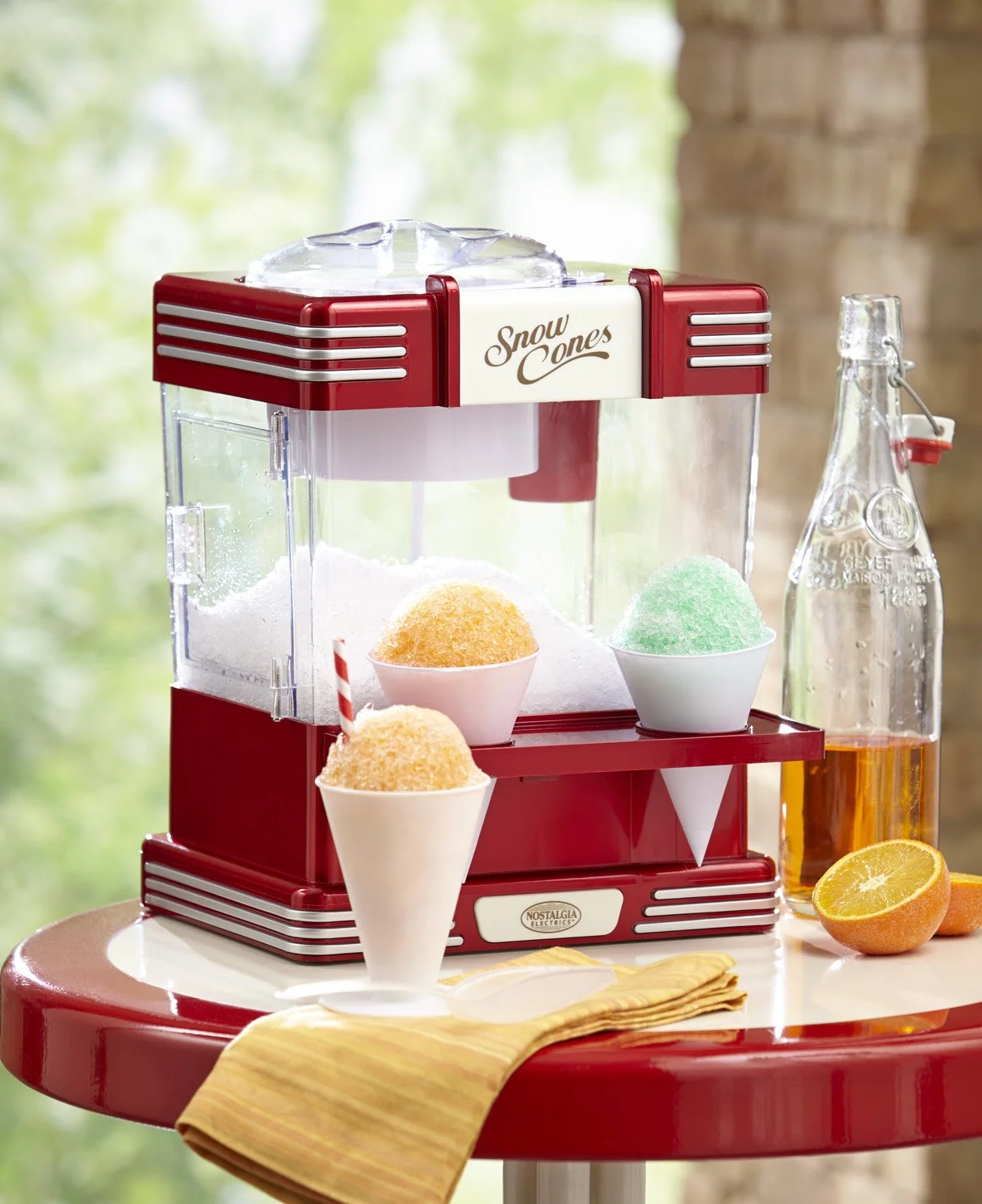 A retro red and white snow cone maker on a table with three colorful snow cones, a bottle of orange soda, and orange halves, with a blurred green outdoor background.