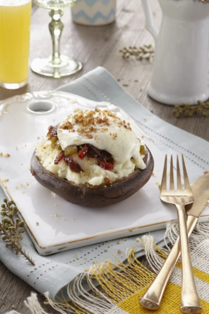 Baked potato topped with sour cream, bacon bits, and chives on a white plate with gold fork and knife, set on a rustic wooden table with a glass of juice, a wine glass, and a white ceramic teapot in the background.