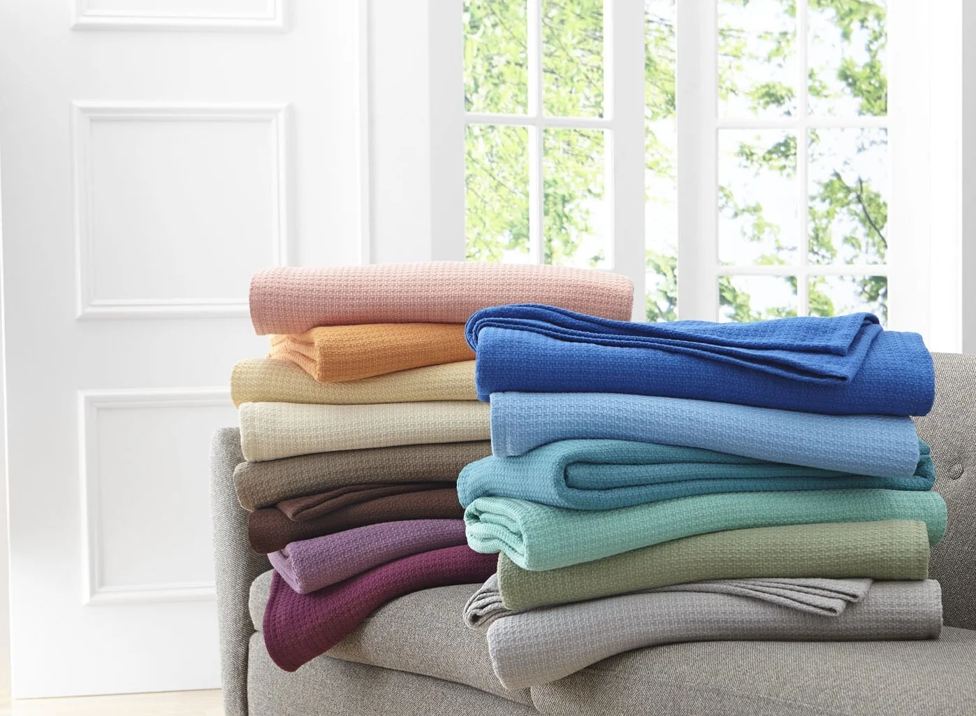 Stack of folded towels in various pastel and bright colors on a light gray sofa, with a large window with white frames and green trees outside in the background.