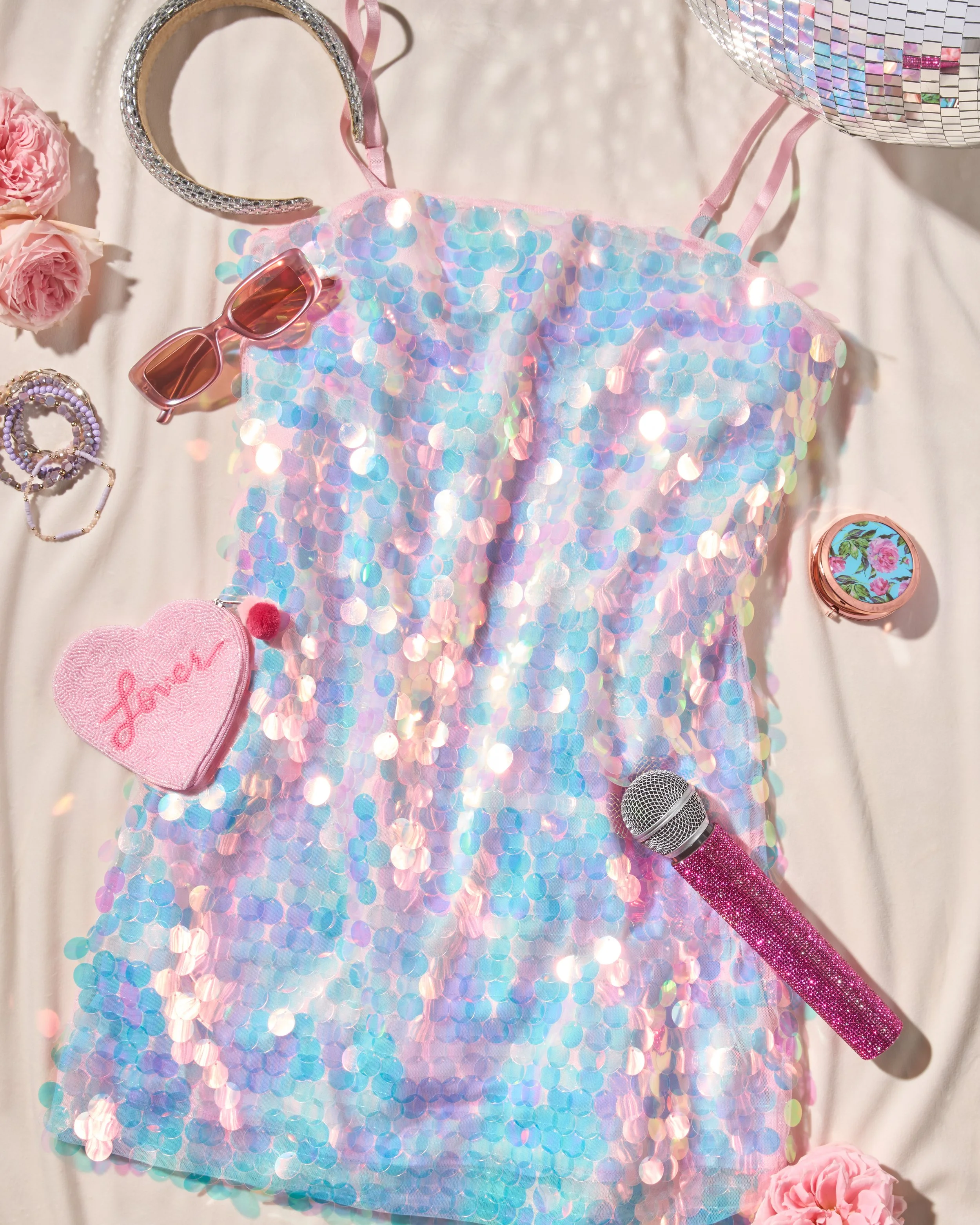 A pink, iridescent sequin dress surrounded by pink roses, sunglasses, jewelry, a pink glittery microphone, and a round compact, all set on a pale surface.