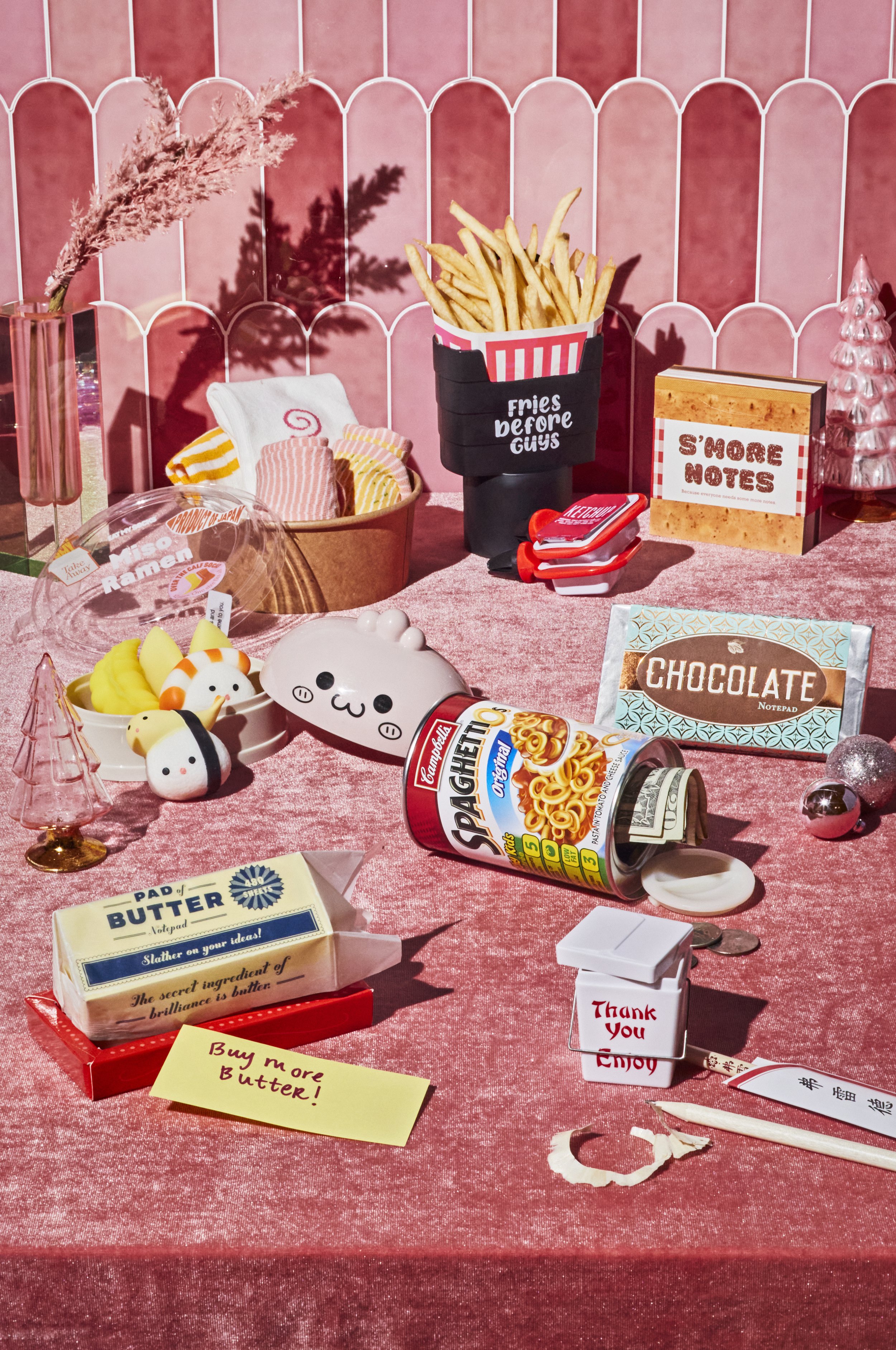A pink-themed table setup with snacks and decorations, including a container of French fries, a can of SpaghettiOs, a notepad labeled "Chocolate," a butter packet with a note saying "Buy more Butter!", various sweets with cute faces, and holiday deco