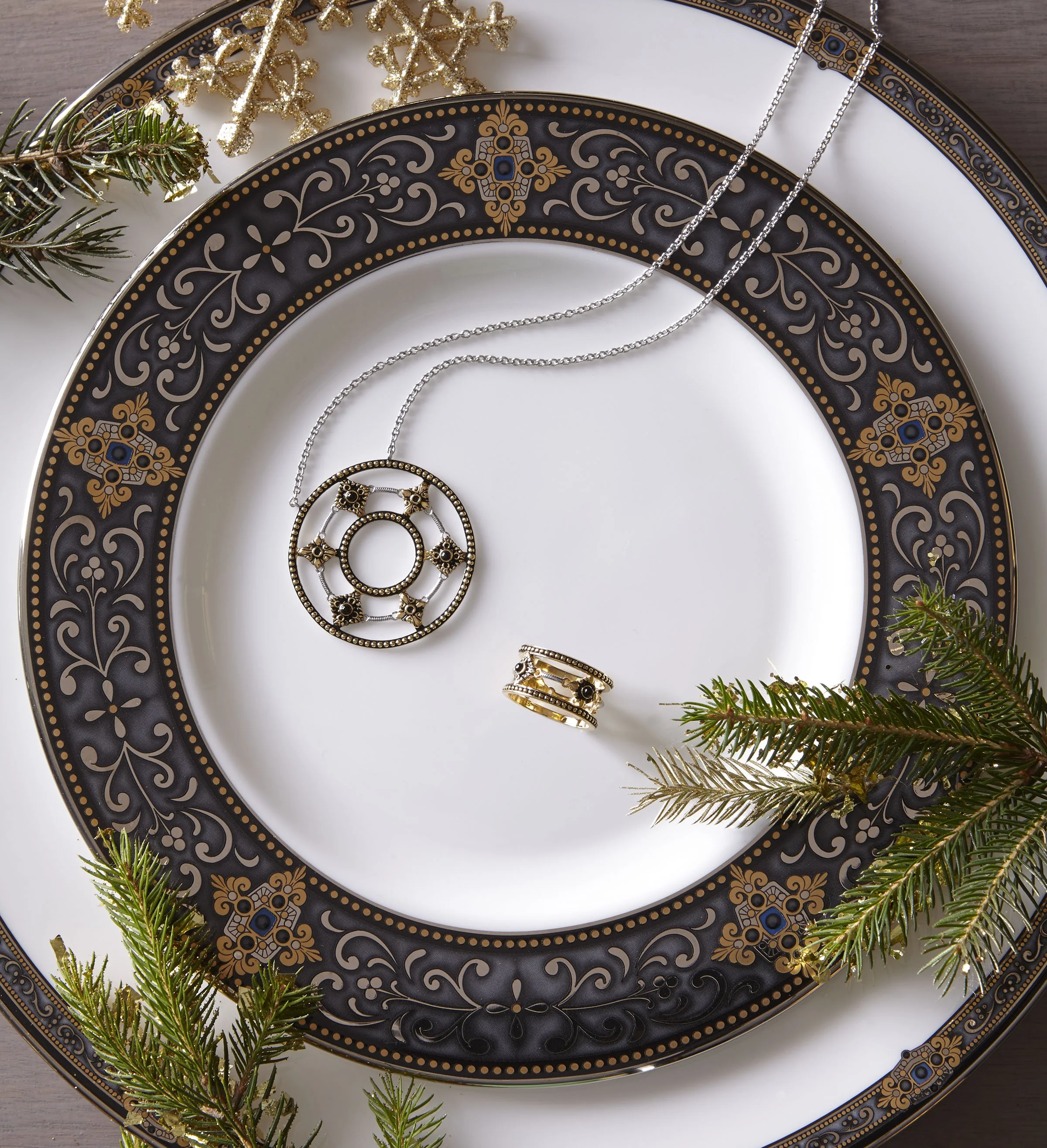 Decorative black and gold patterned plates with holiday evergreen branches, featuring a silver necklace with a round pendant, a gold ring with black stones, and gold earrings with black stones.