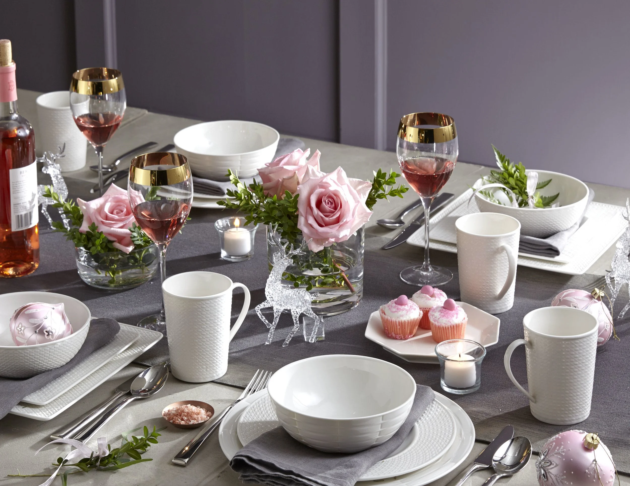 A decorated table set for a celebration with pink roses, cupcakes, pink wine, candles, and Christmas ornaments.