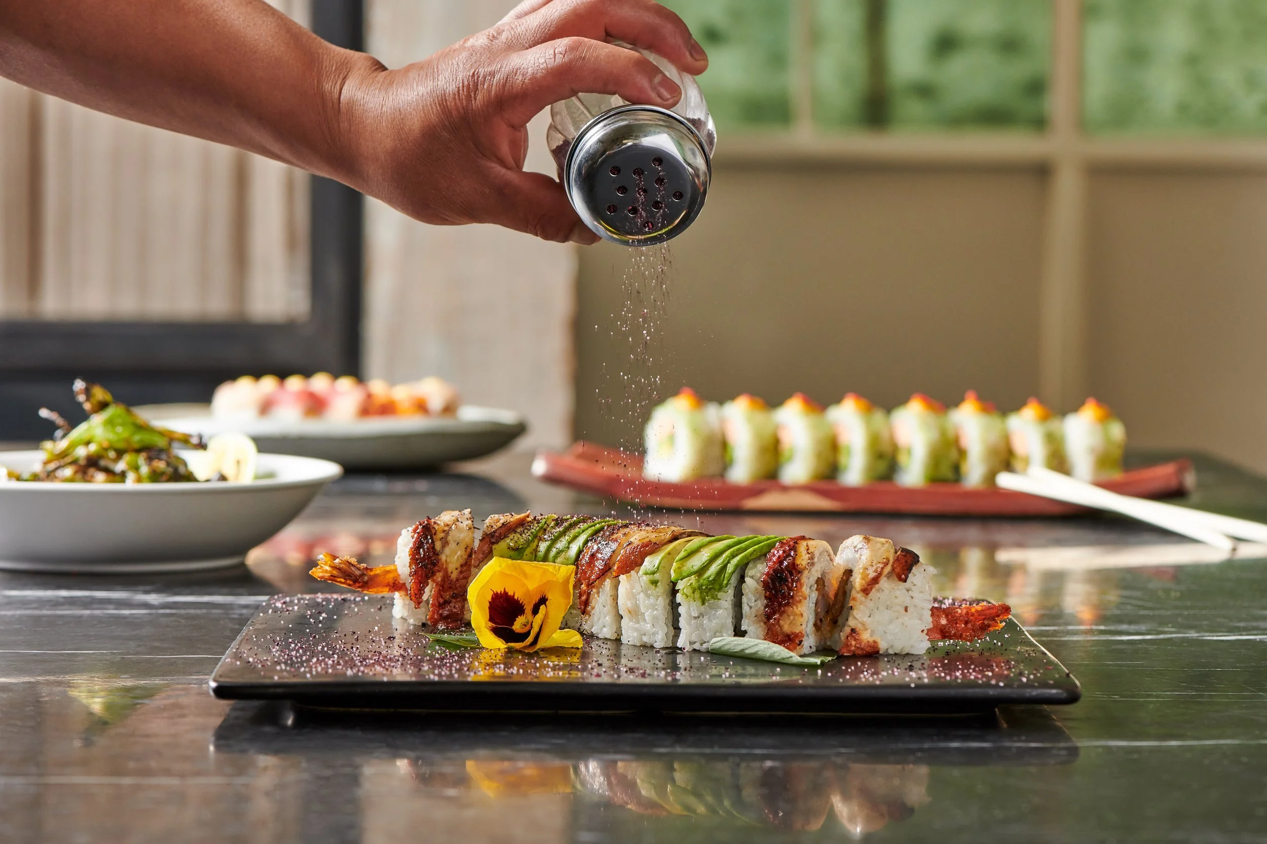 Close-up of a hand sprinkling black pepper on a sushi roll platter, with various sushi rolls and dishes visible in the background.