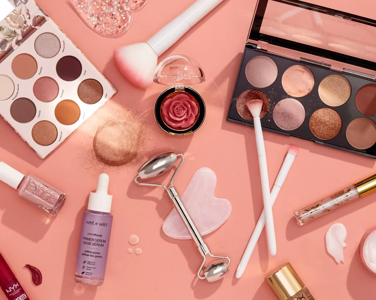 Flat lay of pink and neutrally colored makeup products and tools on a pink surface, including eyeshadow palettes, brushes, skincare bottles, a facial roller, and various cosmetics.