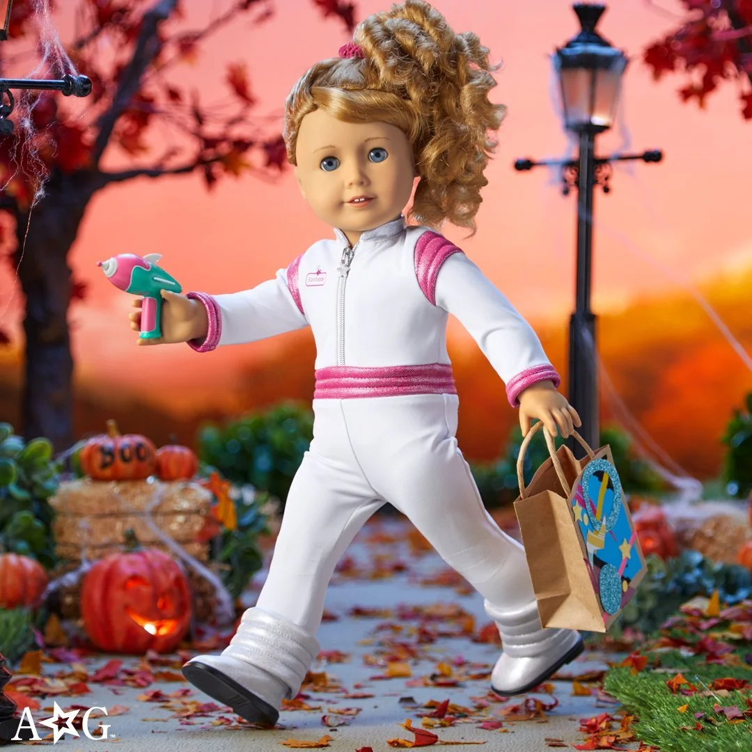 An American Girl doll with curly red hair, dressed in a white and pink tracksuit, holding a toy water gun and a shopping bag, walking through a Halloween-themed outdoor scene with pumpkins, a scarecrow, and fall leaves, during sunset.