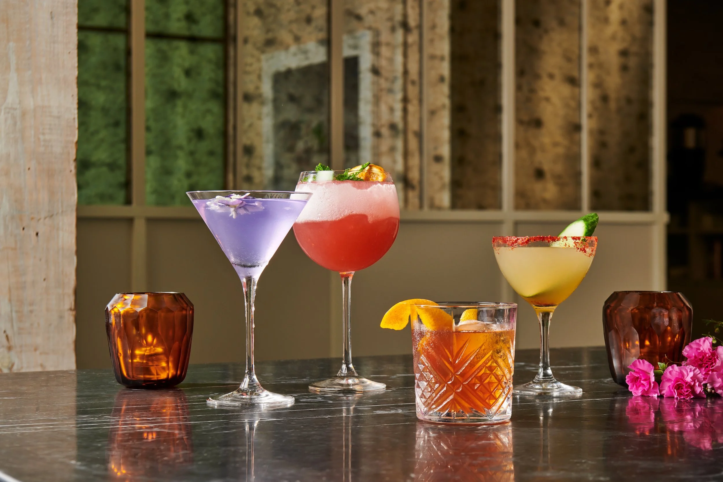 Five colorful cocktails on a dark wooden table, with two amber candles and pink flowers nearby, set against a backdrop of large windows and brick walls.