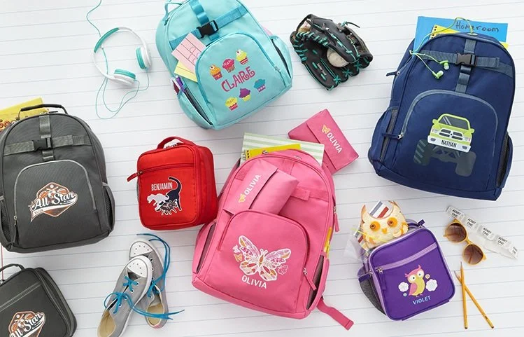 School supplies including backpacks, notebooks, sunglasses, headphones, and small accessories arranged on a white lined surface.