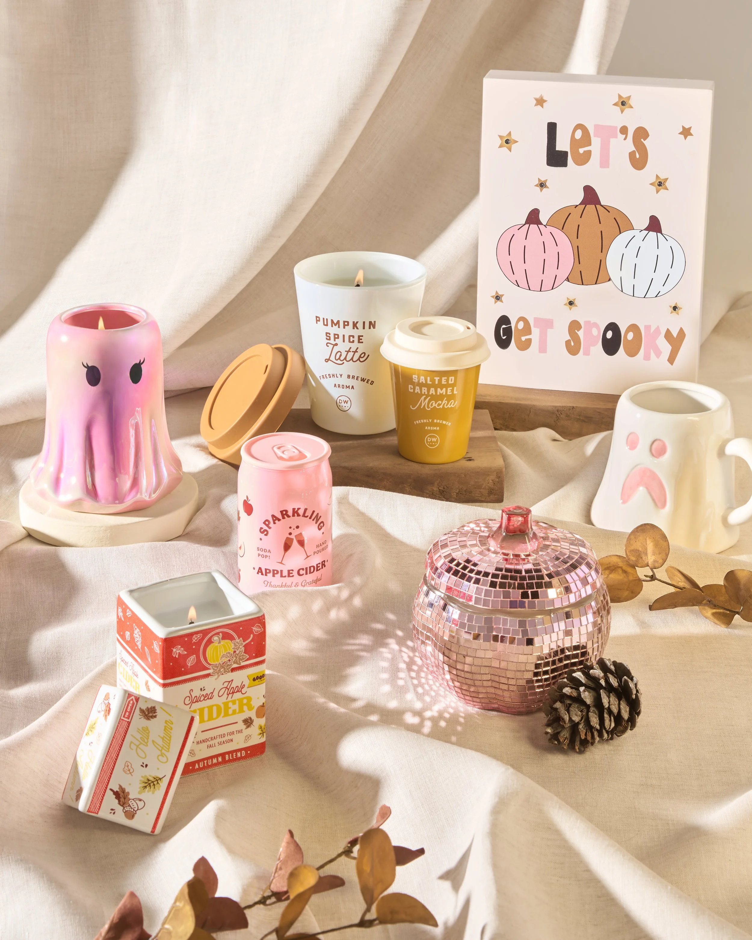 Decorative autumn-themed candles, ceramic mugs, and a pink disco ball are arranged on a cream-colored fabric with a "Let's Get Spooky" sign featuring pumpkins in the background.