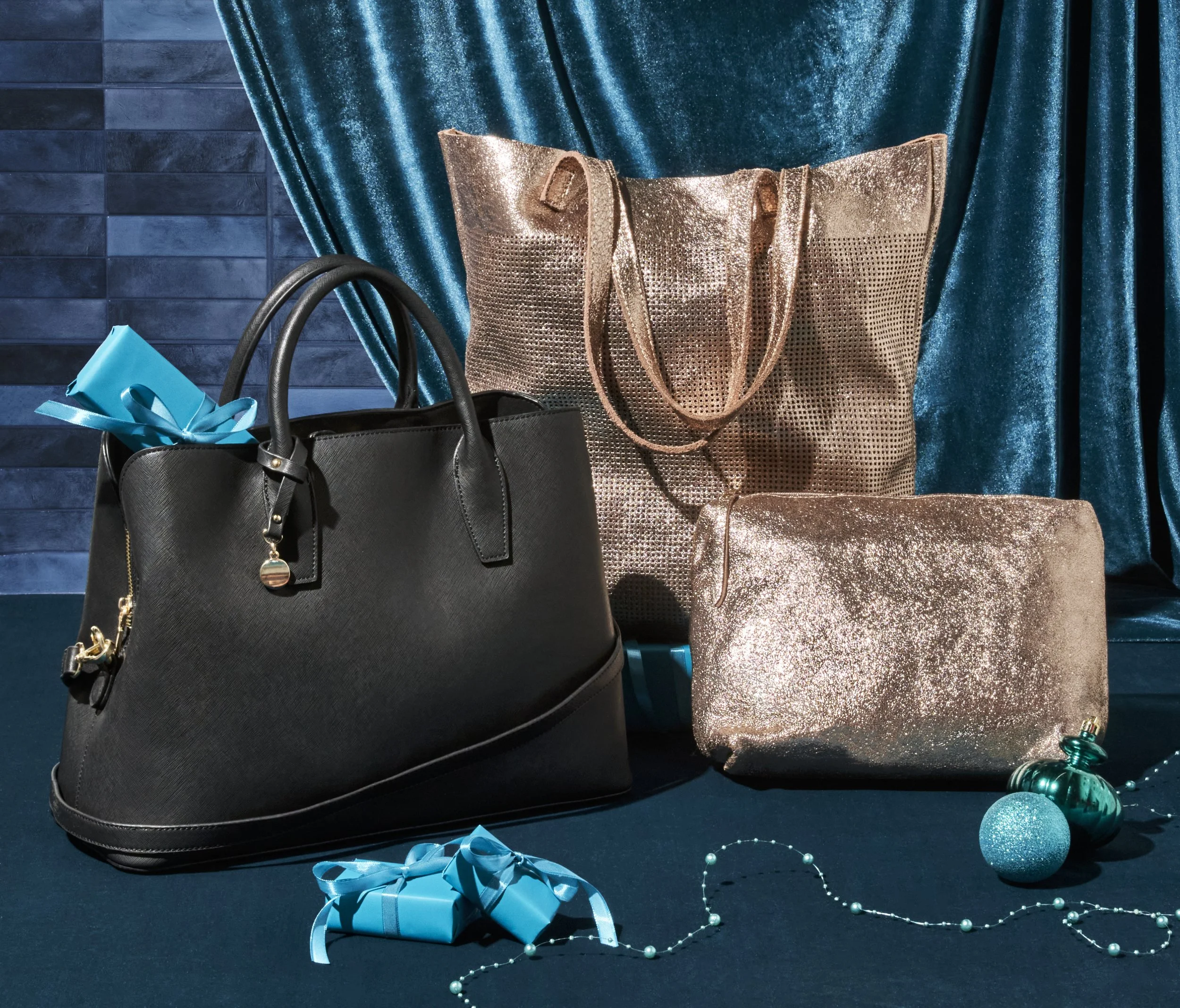 Black handbag, gold sequin tote, and gold sequin clutch fairy tale with blue gift box, blue ribbon, pearl necklace, and Christmas ornaments in front, against a blue curtain backdrop.