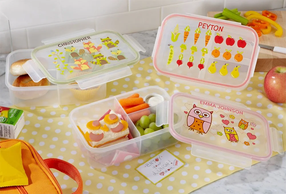 Various colorful personalized kids' lunch boxes with cartoon characters and vegetables on a yellow polka dot tablecloth, surrounded by food and a cutting board with cut vegetables.