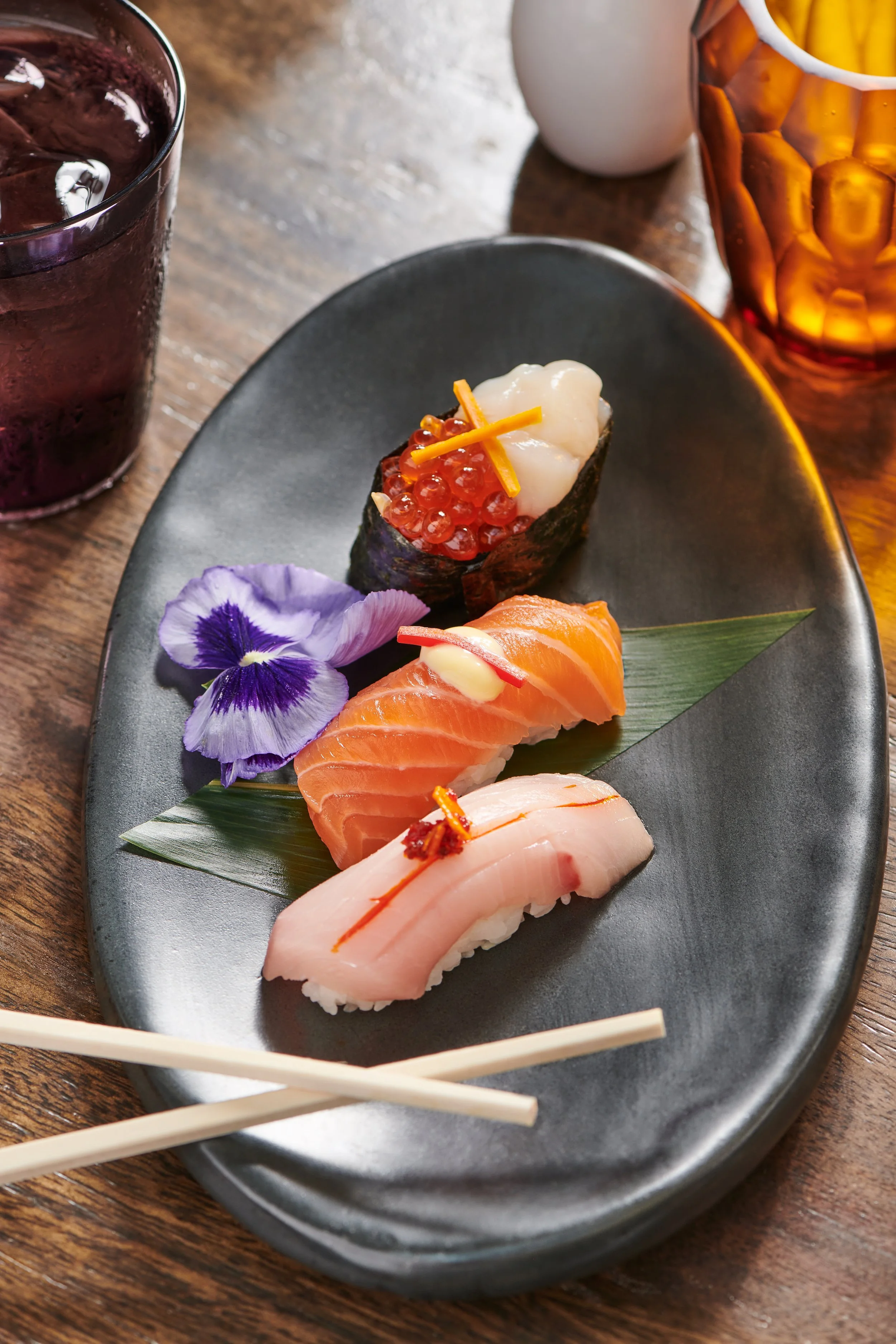 Sushi platter with three pieces of nigiri sushi topped with fish, a flower, and a sushi roll with fish roe, on a black ceramic plate, with a purple drink and a glass of iced tea nearby.