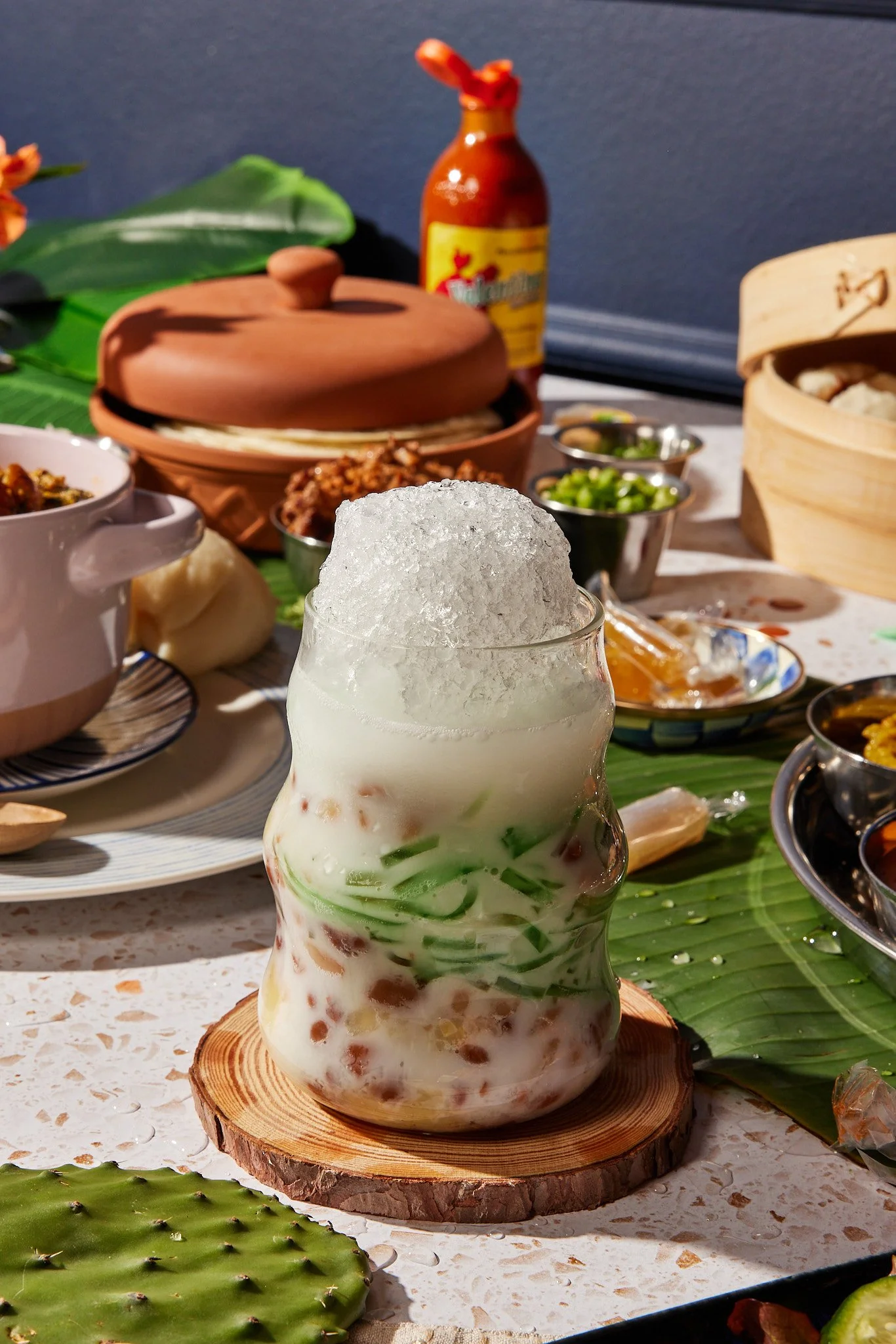A glass of Chinese herbal tea with crushed ice on top, surrounded by various Asian dishes and condiments on a table decorated with banana and cactus leaves.