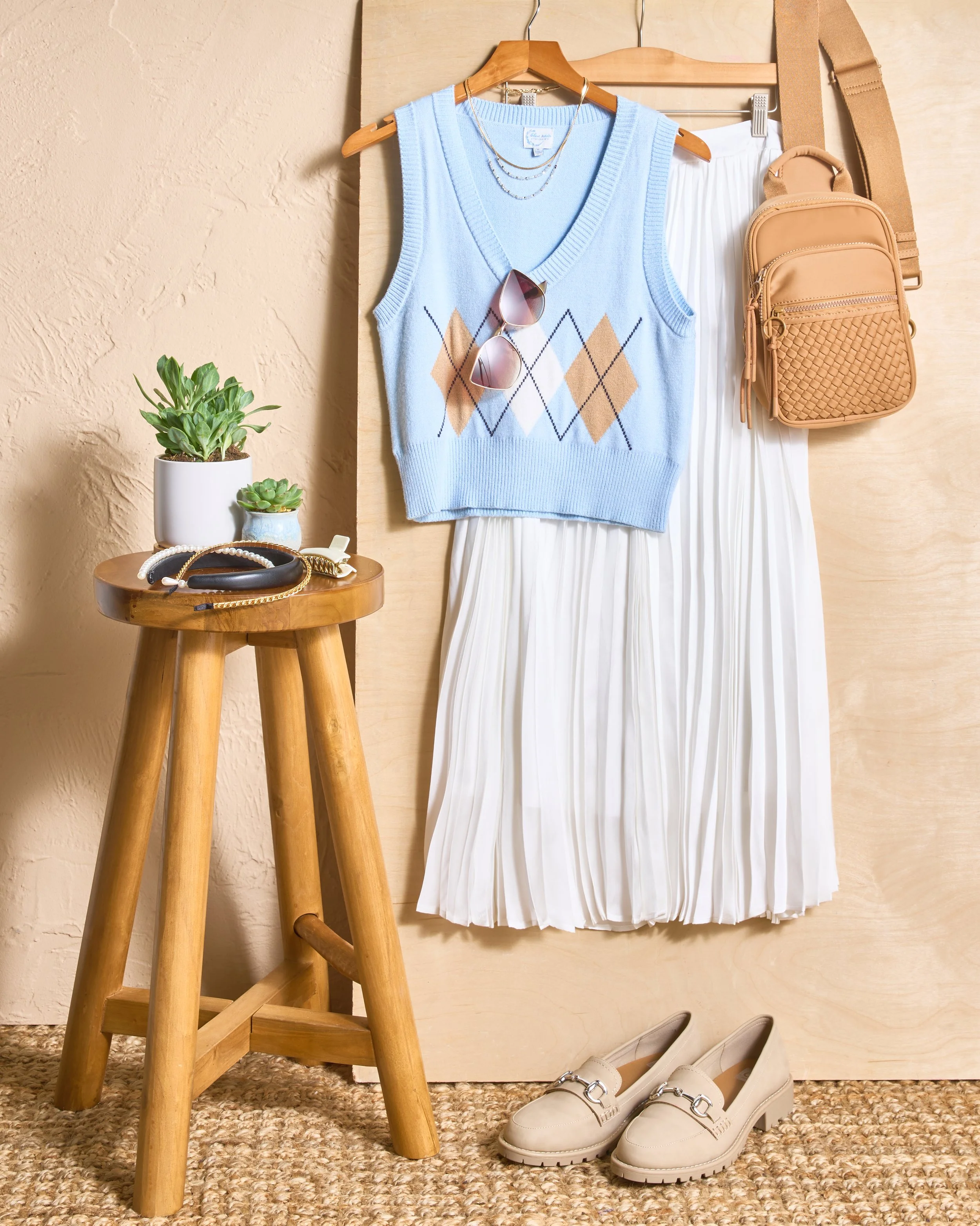 A display of women's fashion items including a light blue argyle sweater vest, white pleated skirt, beige loafers, sunglasses, layered necklaces, and a tan backpack, arranged on a wooden background with a small wooden stool holding plants and jewelry