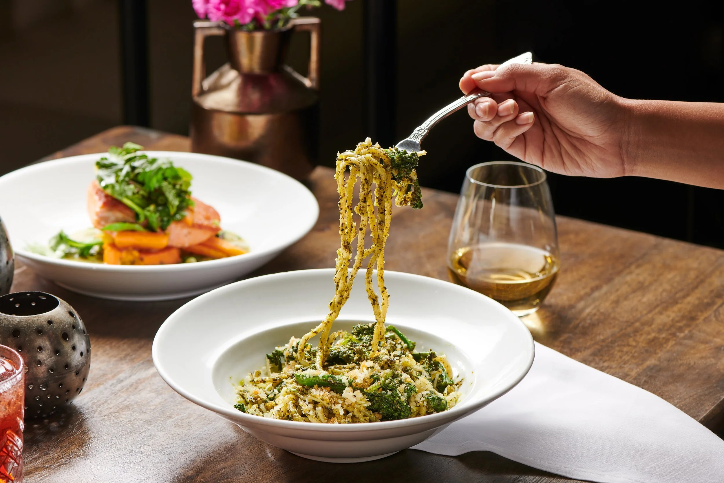 Person lifting a forkful of green pasta from a white bowl on a wooden table, with a glass of white wine and a plate of salad in the background.