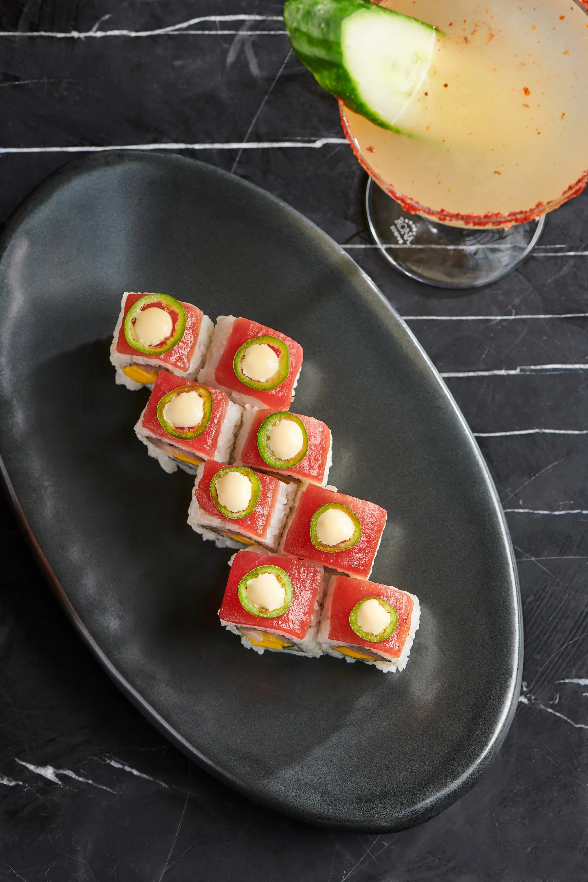 A black oval plate with eight pieces of sushi topped with slices of red fish and green chili rings, served on a dark slate surface with a glass of cocktail garnished with a cucumber slice in the background.