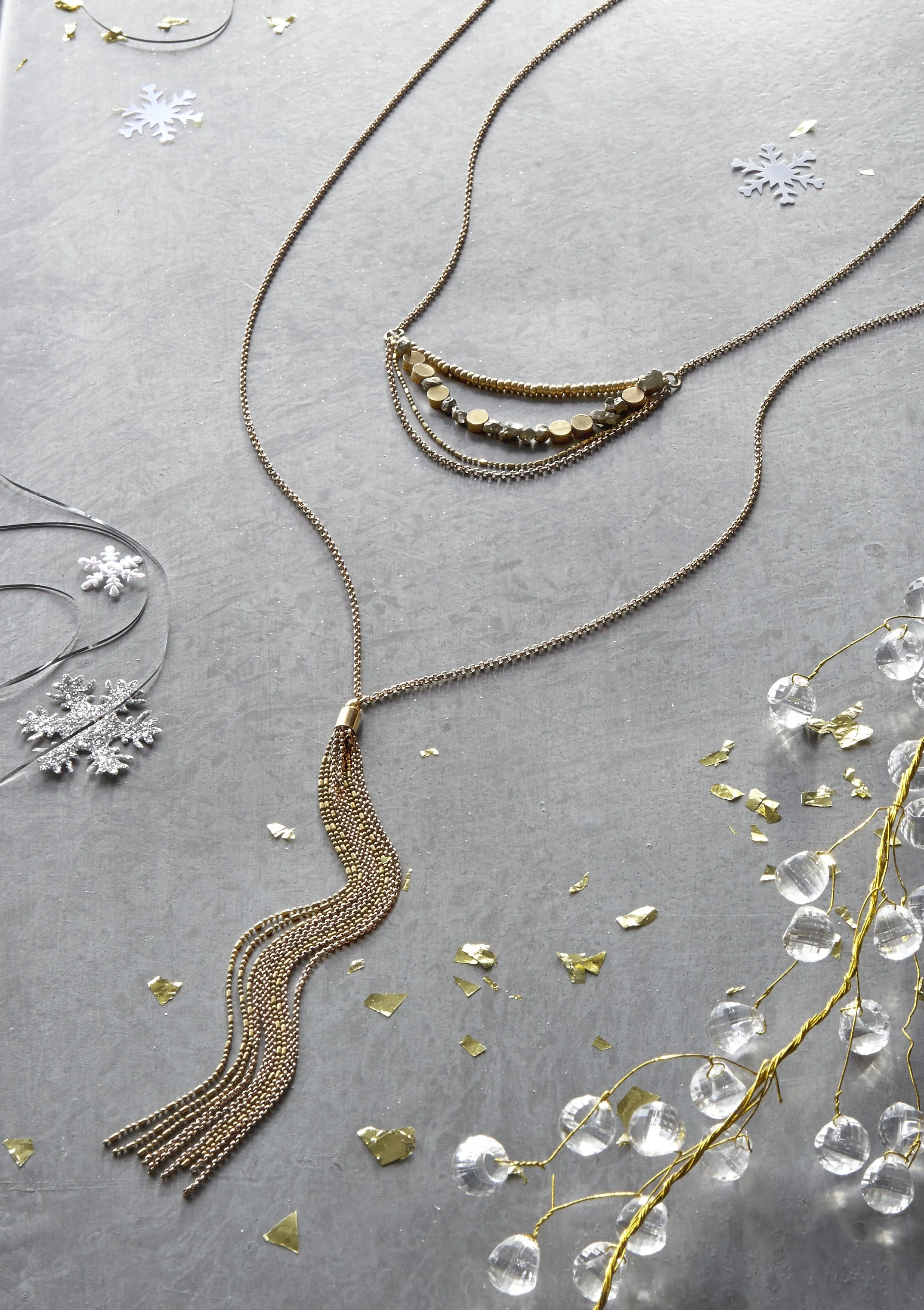 Three layered necklaces with gold and silver beads and chains, surrounded by snowflake and glitter decorations on a gray background.