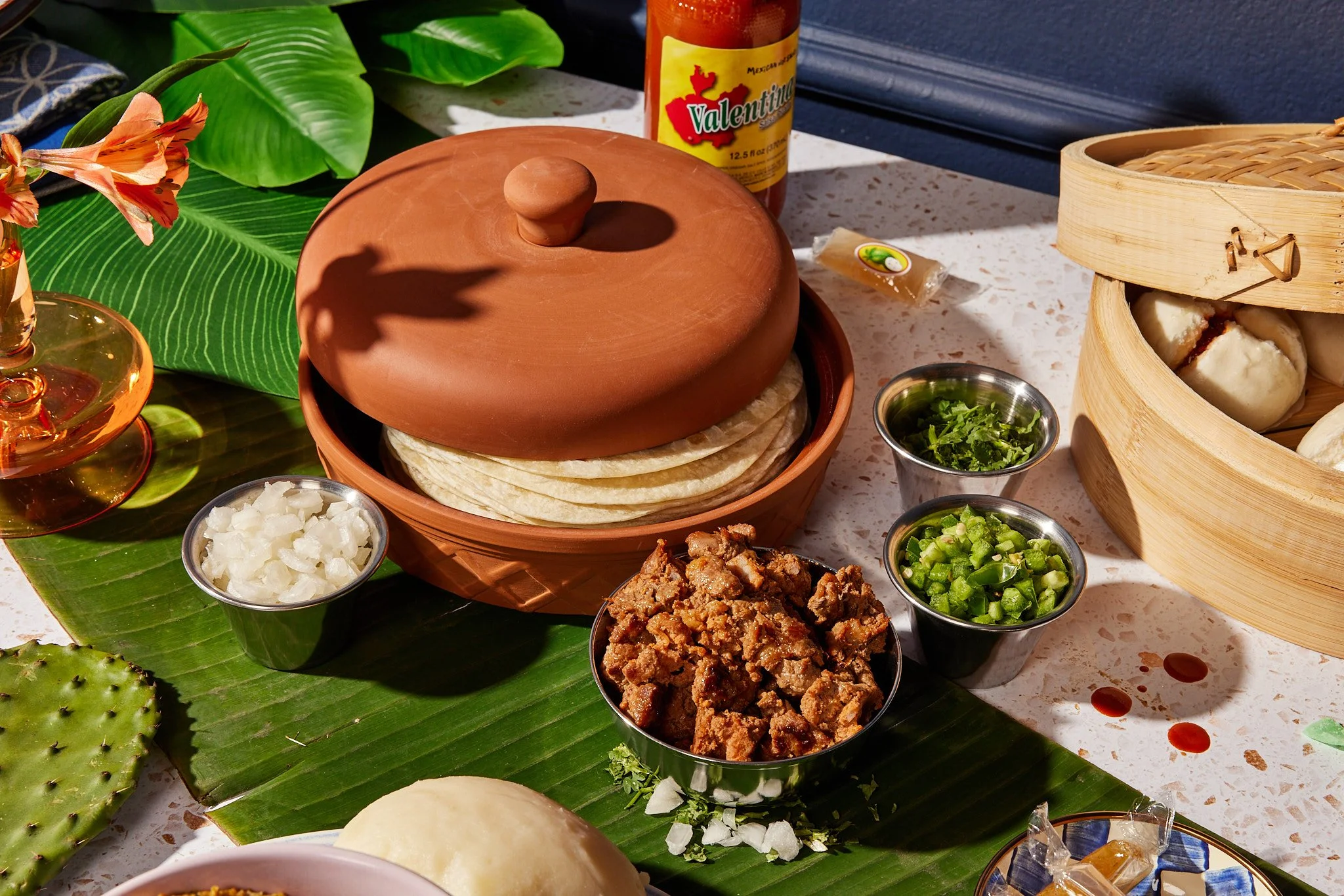 A traditional Mexican food spread with a clay tortilla warmer holding tortillas, accompanied by small bowls of chopped onions, cilantro, and jalapeños on a banana leaf, with a basket of tamales and a bottle of Valentina hot sauce.