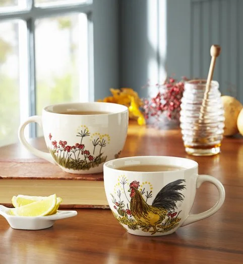 Two decorative teacups with floral and rooster designs on a wooden table, with lemon wedges, honey jar, and a window in the background.