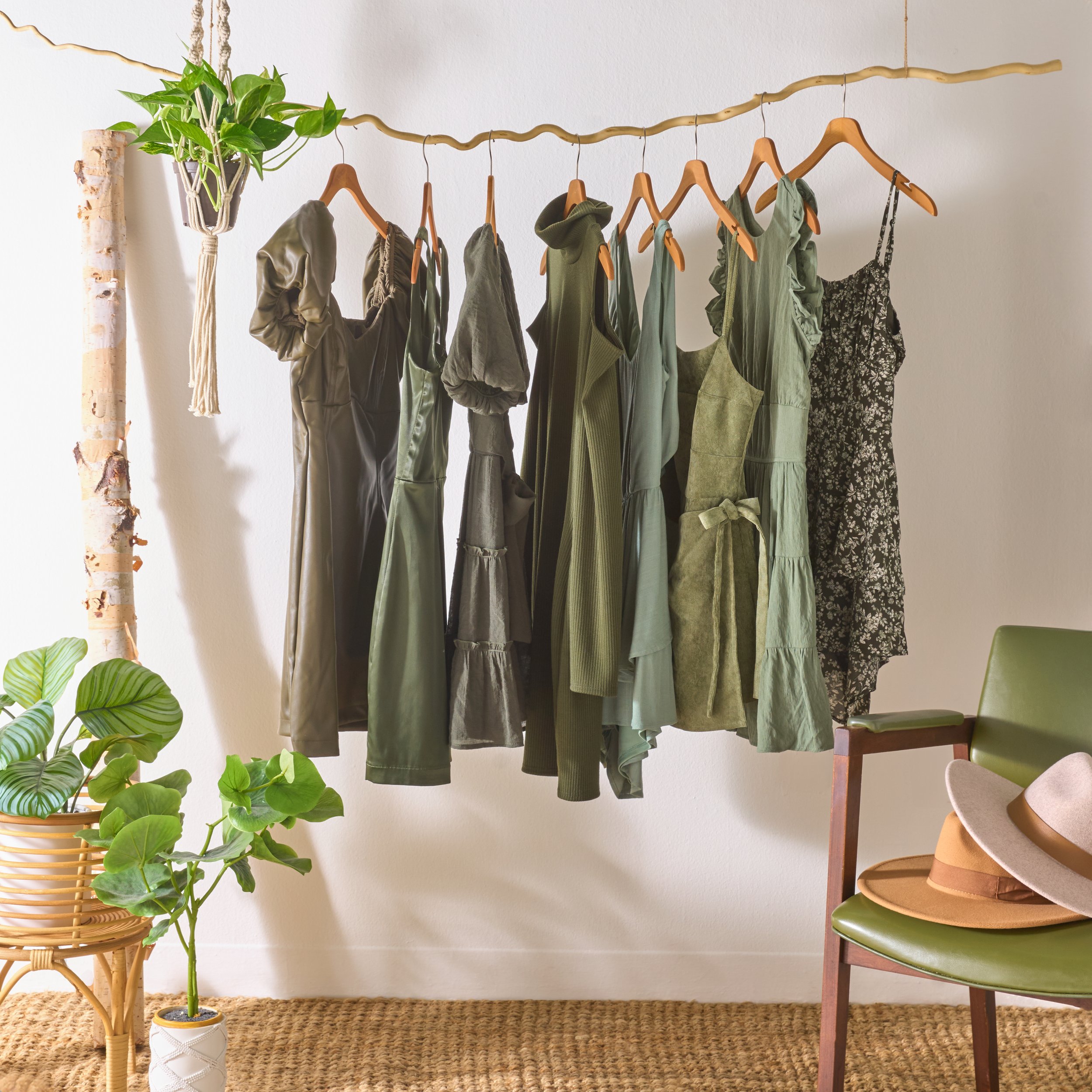 A clothing rack with various green and black dresses and garments hanging on wooden hangers, with houseplants and a hat on a chair nearby.