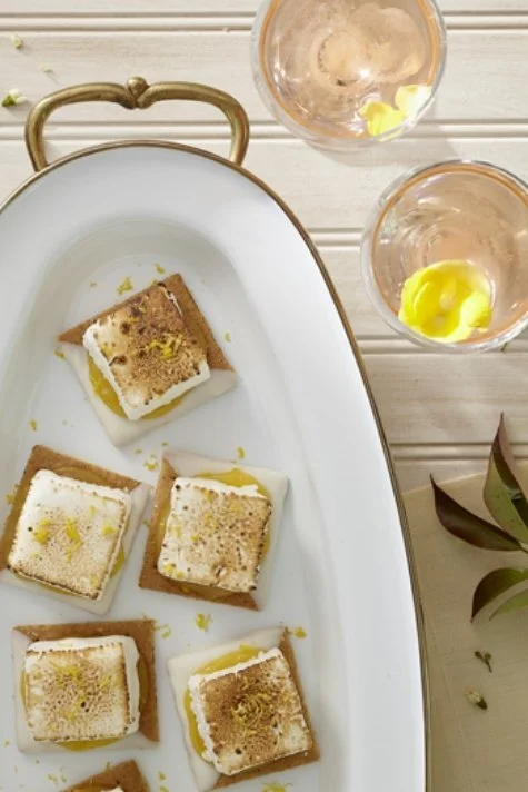A white plate with square crackers topped with melted white cheese, lemon zest, and black pepper, with two glasses of water with lemon slices nearby.