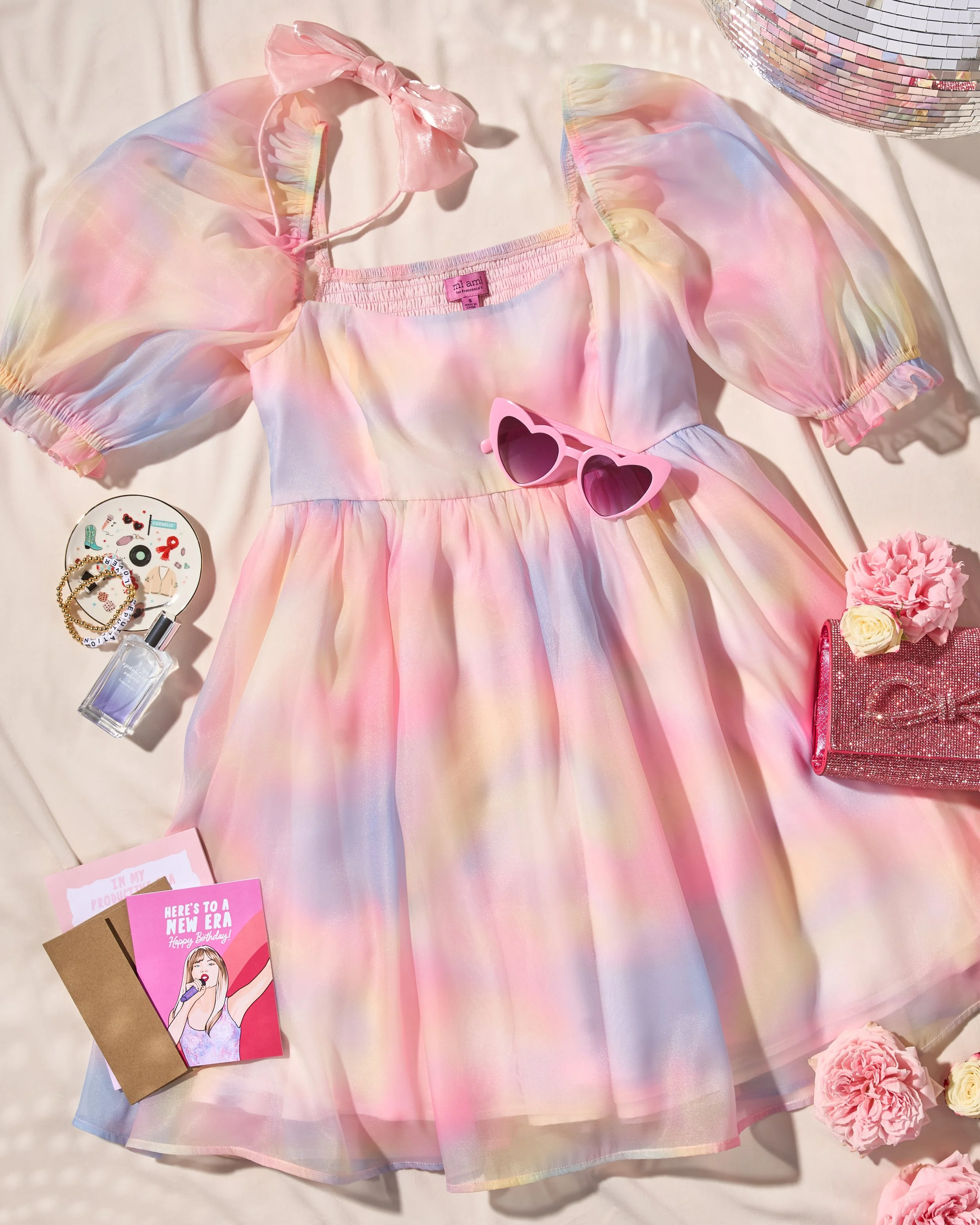 A pastel-colored, iridescent dress with puffed sleeves and a pink bow on one shoulder, pink heart-shaped sunglasses, a pink clutch, a bottle of perfume, jewelry, greeting cards, and pink flowers arranged on a light background.