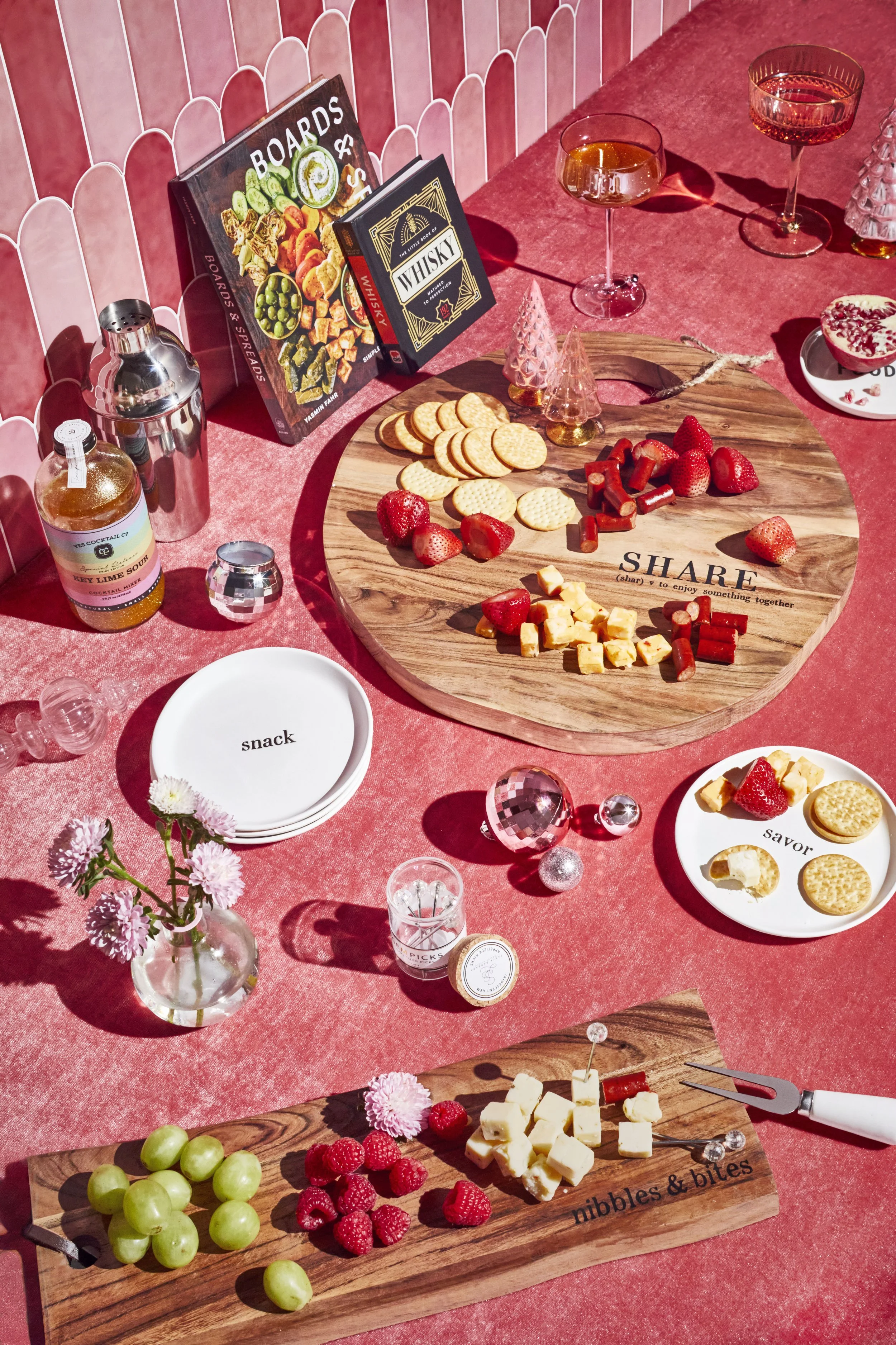 An overhead view of a table set with cheese, crackers, strawberries, raspberries, grapes, and festive decorations including pink and silver ornaments, a small glass vase with pink flowers, and books, under pink wallpaper.