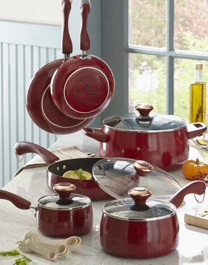 Red cookware set on a kitchen table with pots, pans, and hanging pans near a window.